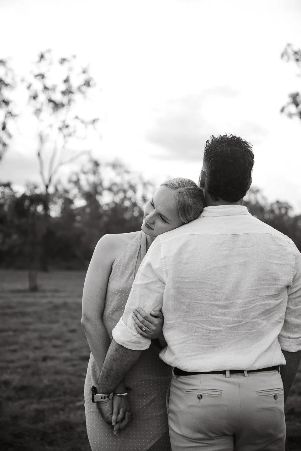 A woman with long hair resting her head on a man's shoulder, holding his hand, outdoors in a park setting, black and white photo.