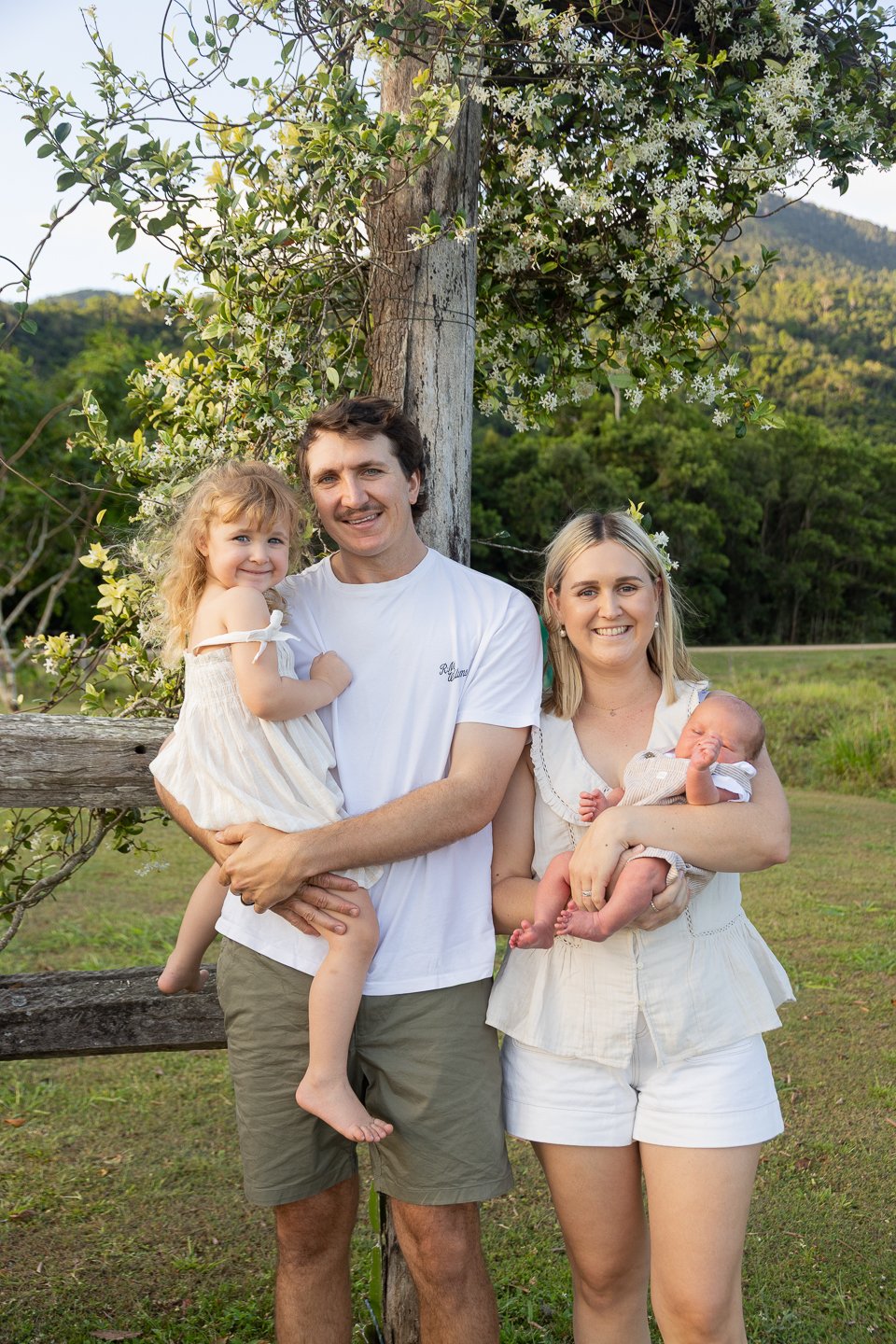 A family of four, including a man, a woman, a young girl, and a newborn baby, standing outdoors in front of a tree and greenery, smiling at the camera.