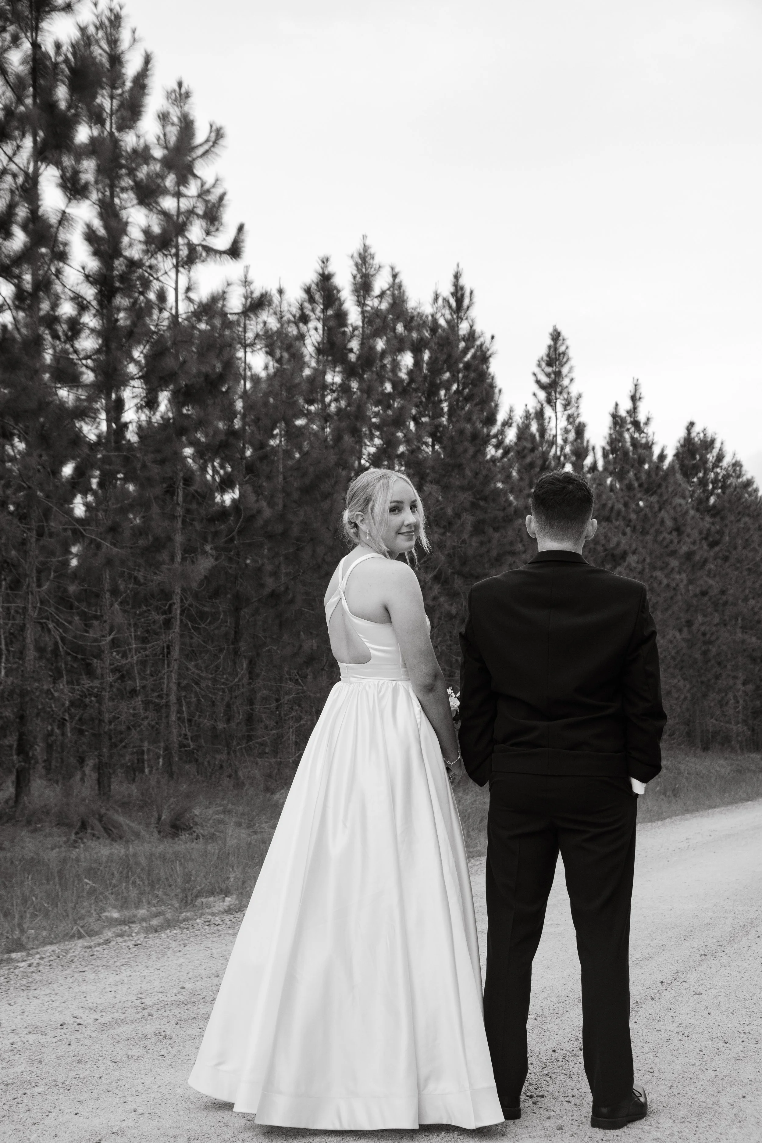 A bride and groom holding hands on a dirt road with a forest in the background, black and white photo.