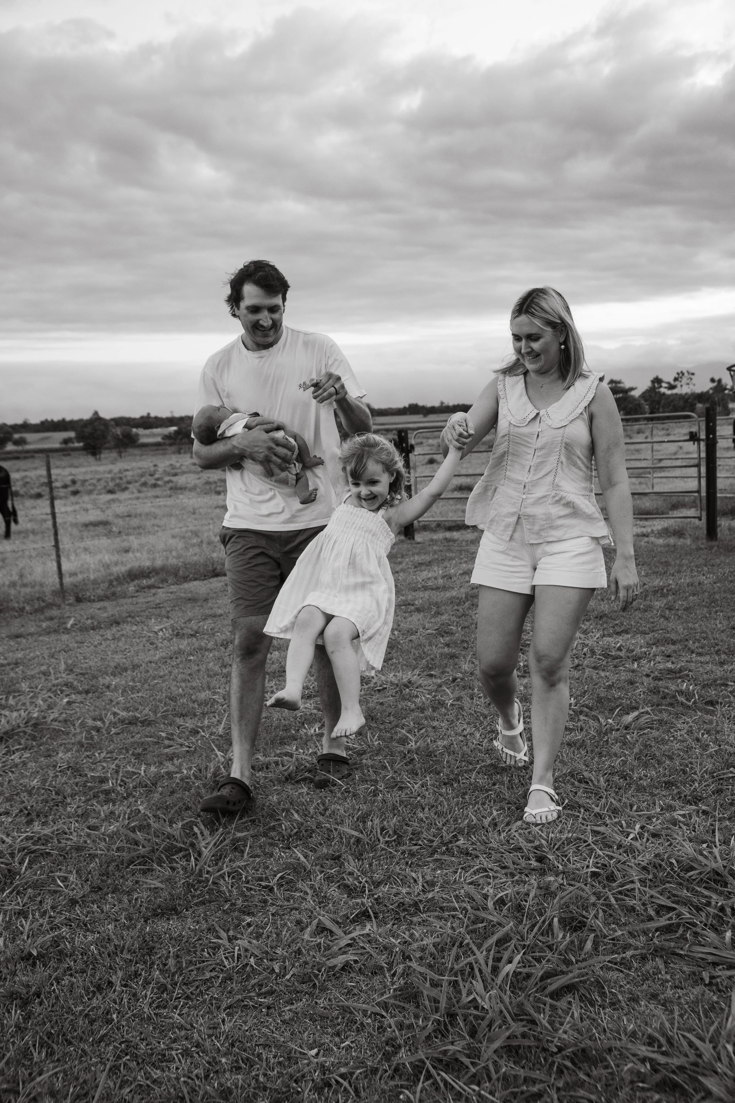 A family of four walking on a grassy field outdoors, with a man carrying a baby, a woman holding hands with a young girl, and an open farm with fences and trees in the background under cloudy skies.