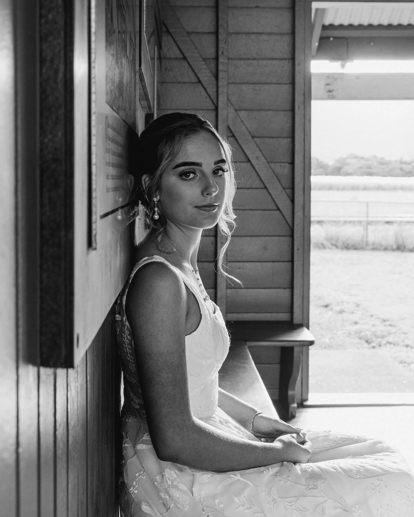 A young woman in a wedding dress sitting inside a wooden structure, looking at the camera, with a scenic outdoor view in the background.
