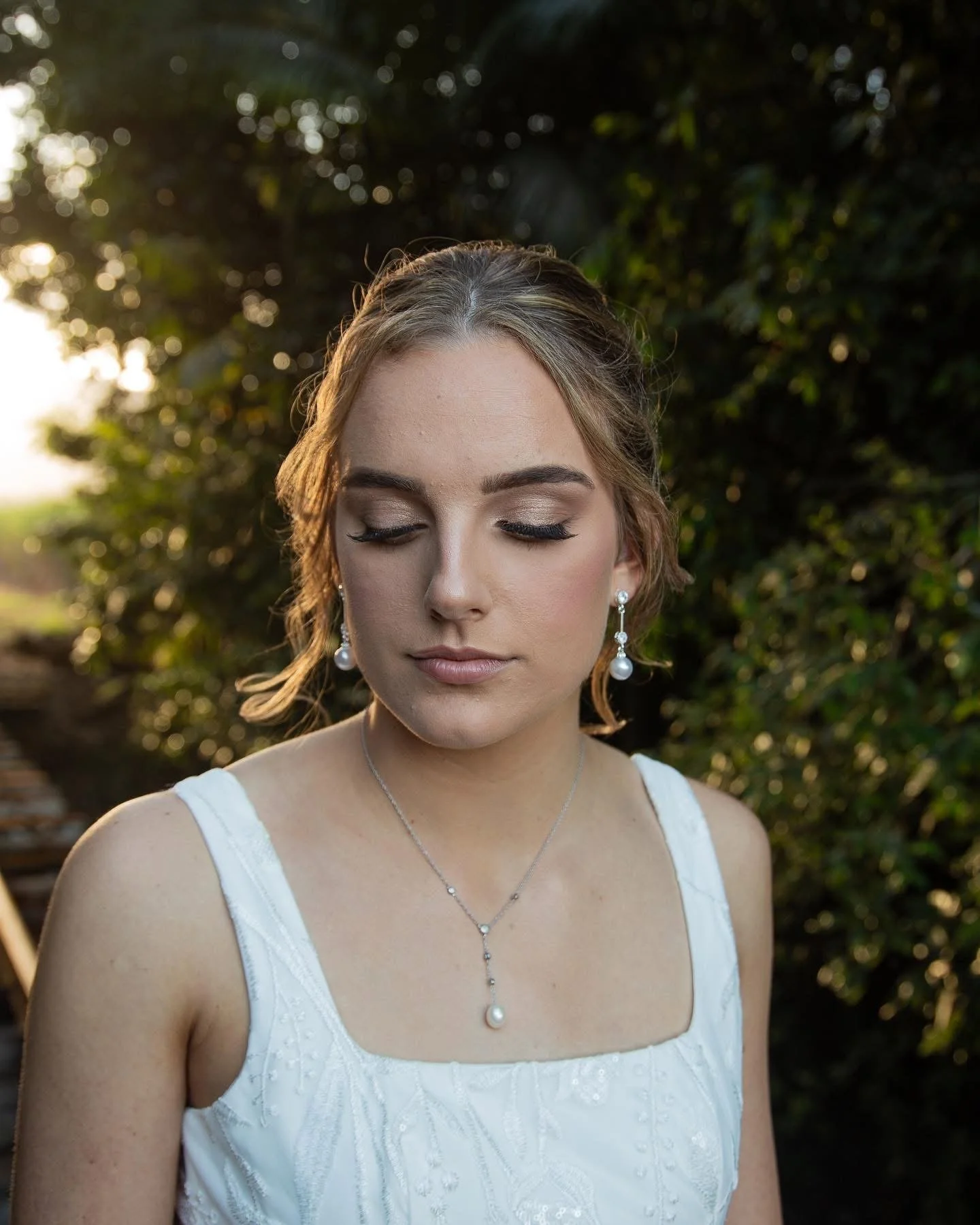 A young woman with makeup, wearing pearl earrings and a pearl necklace with a teardrop pendant, closed eyes, and a white dress, outdoors with greenery and sunlight in the background.
