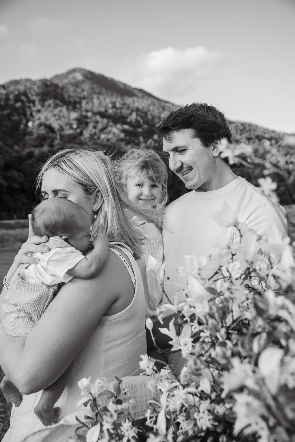 A black-and-white photo of a family outdoors with mountain scenery in the background. A woman is holding a baby, a young girl is smiling, and a man is standing nearby. Flowers are in the foreground.