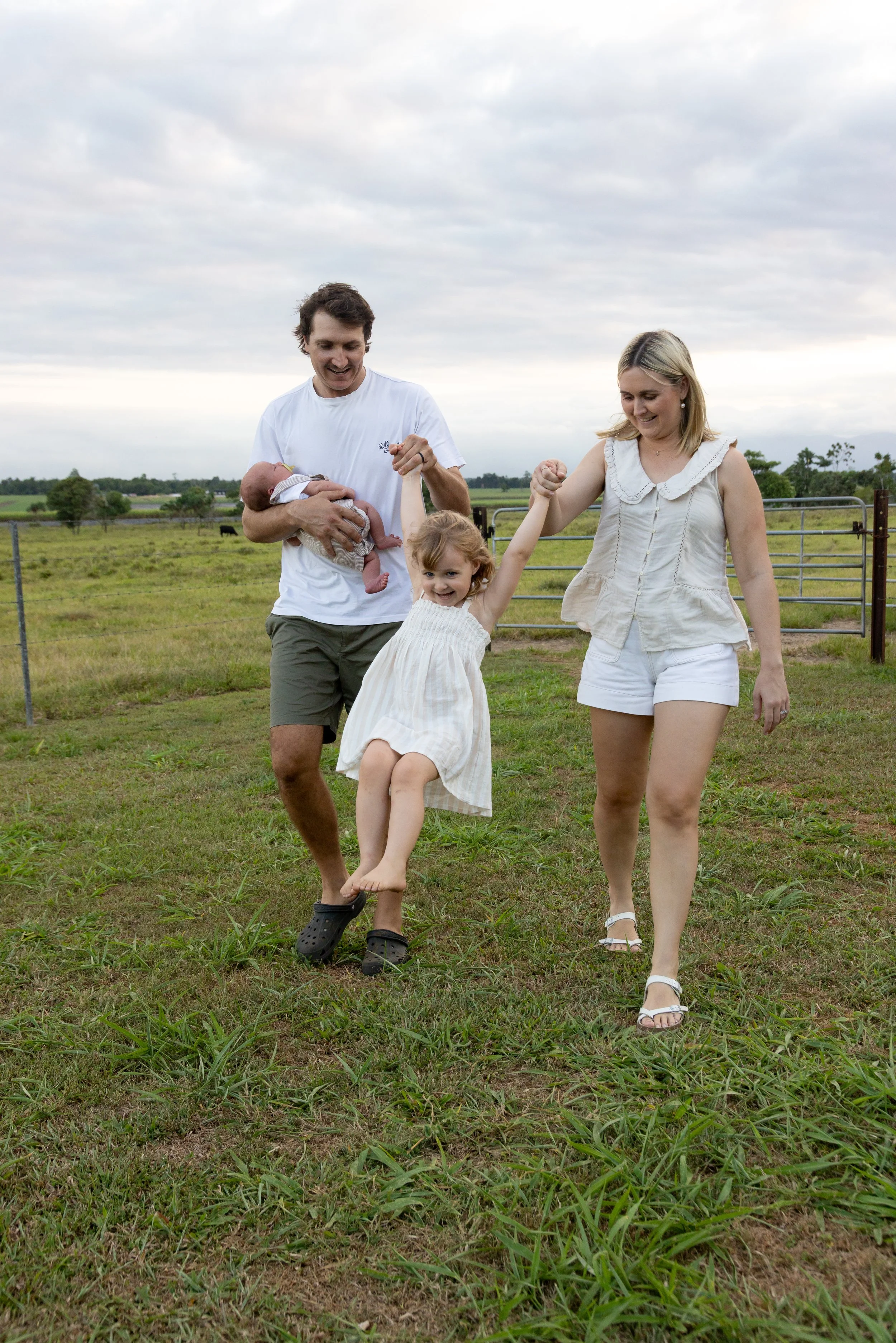Family of four on a farm: father holding newborn, mother and daughter playing on a grassy field.
