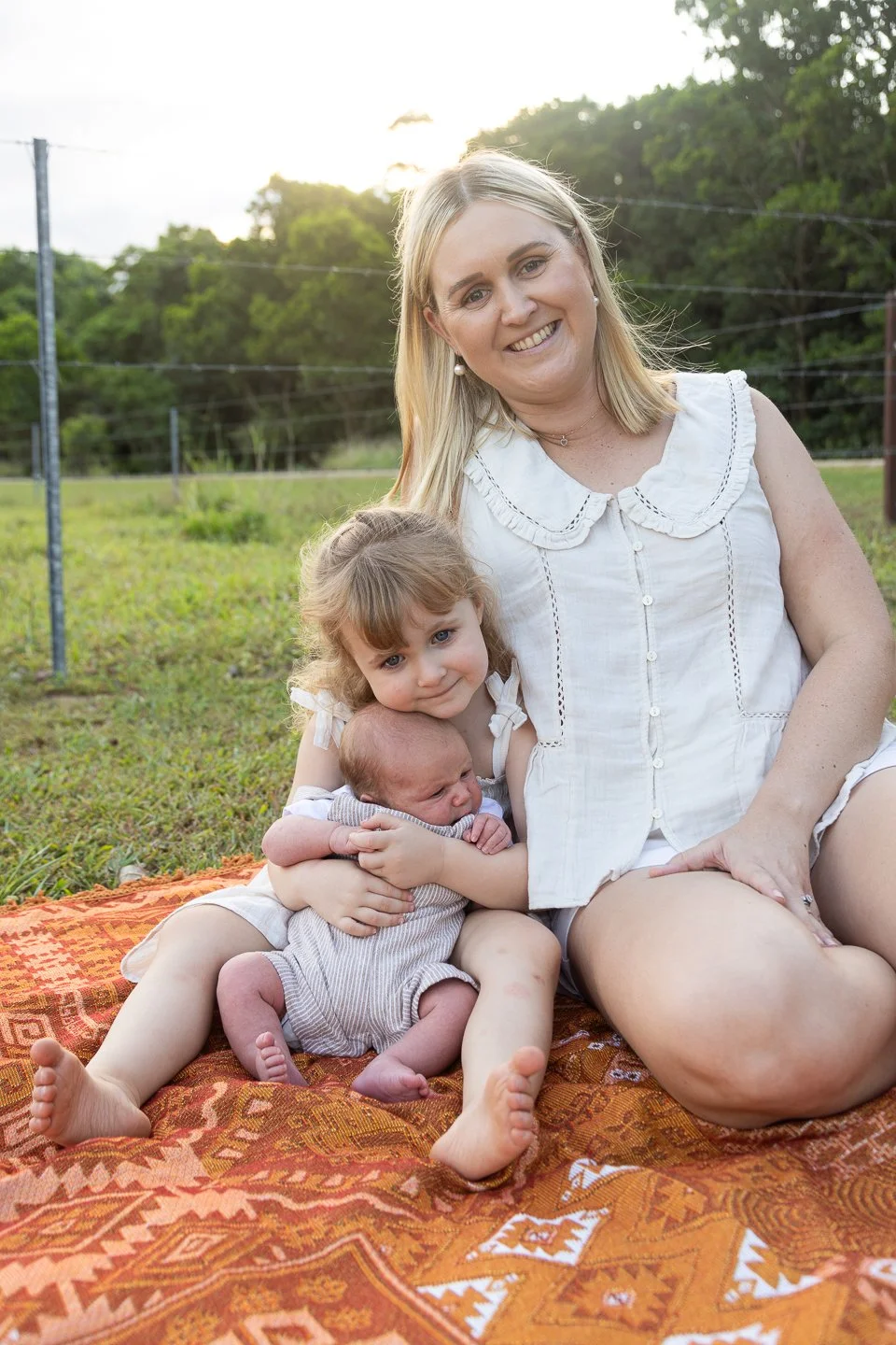 A woman sitting outdoors on a patterned orange blanket, holding two children, with a green, wooded background and sunlight in the sky.