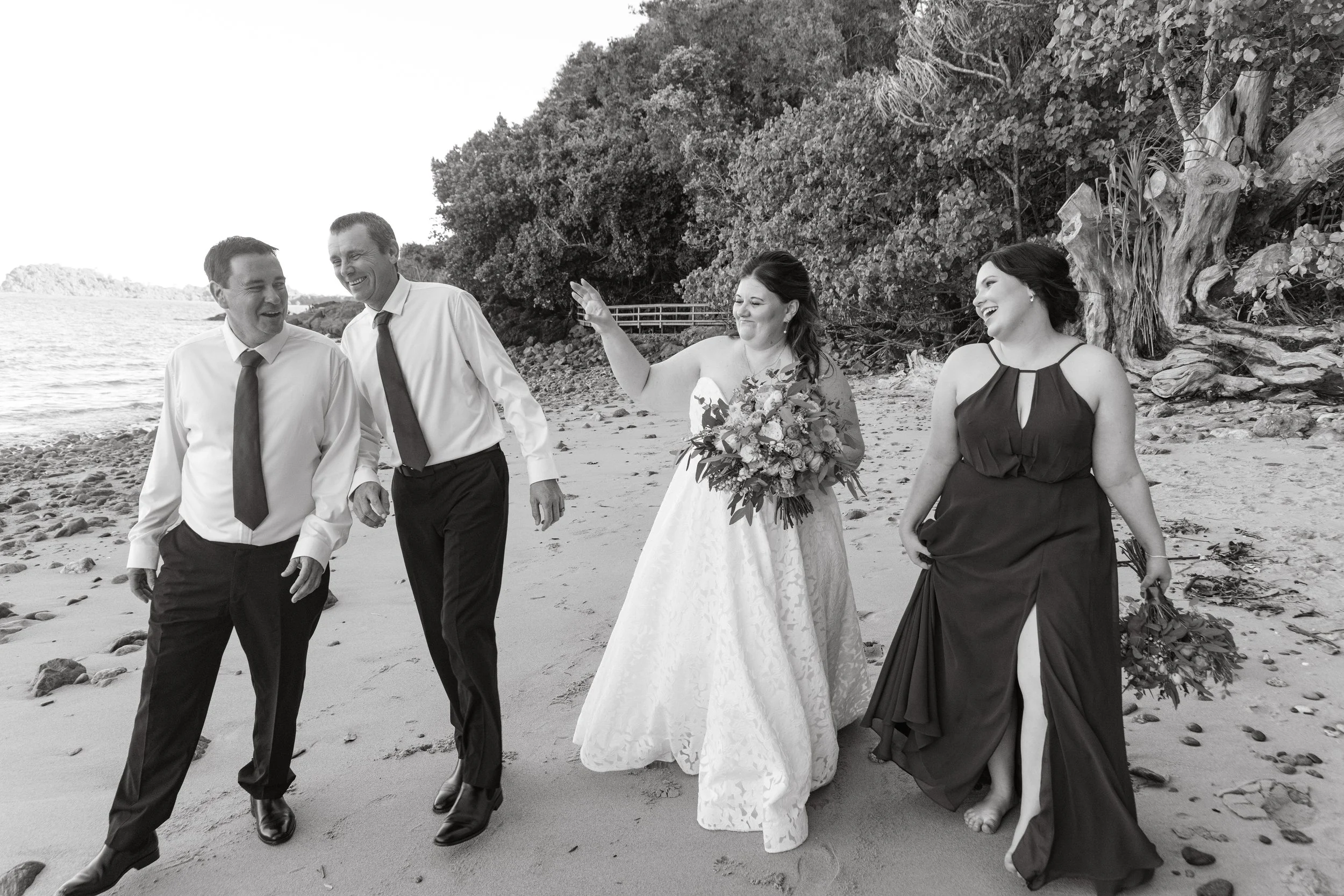 Black and white photo of a wedding party walking along a beach, with two men in white shirts and dark pants, and two women in elegant dresses holding bouquets, all smiling and enjoying their time.
