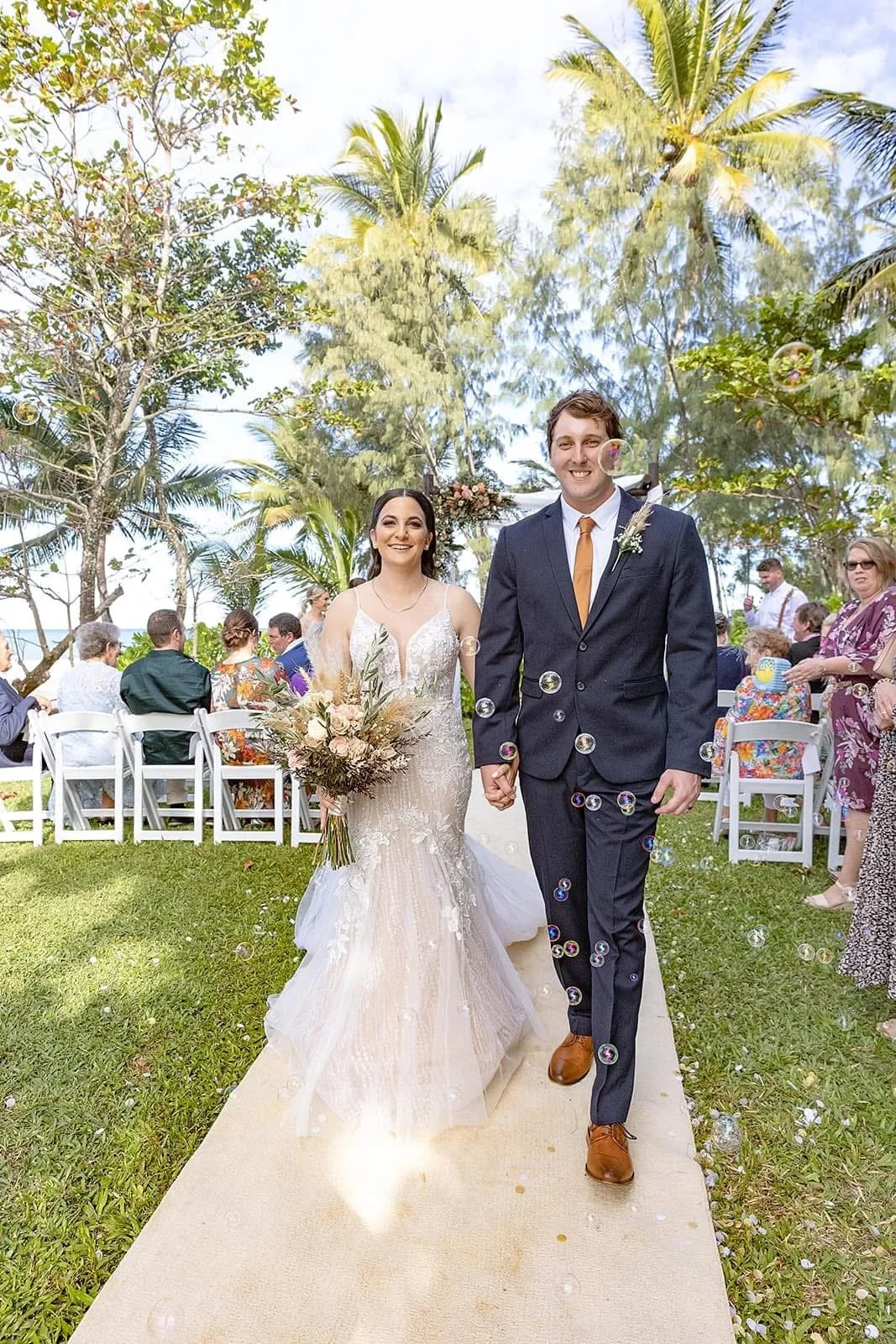 A bride and groom walking hand-in-hand outdoors during their wedding ceremony, surrounded by guests and palm trees, with bubbles floating in the air and a bright sky.
