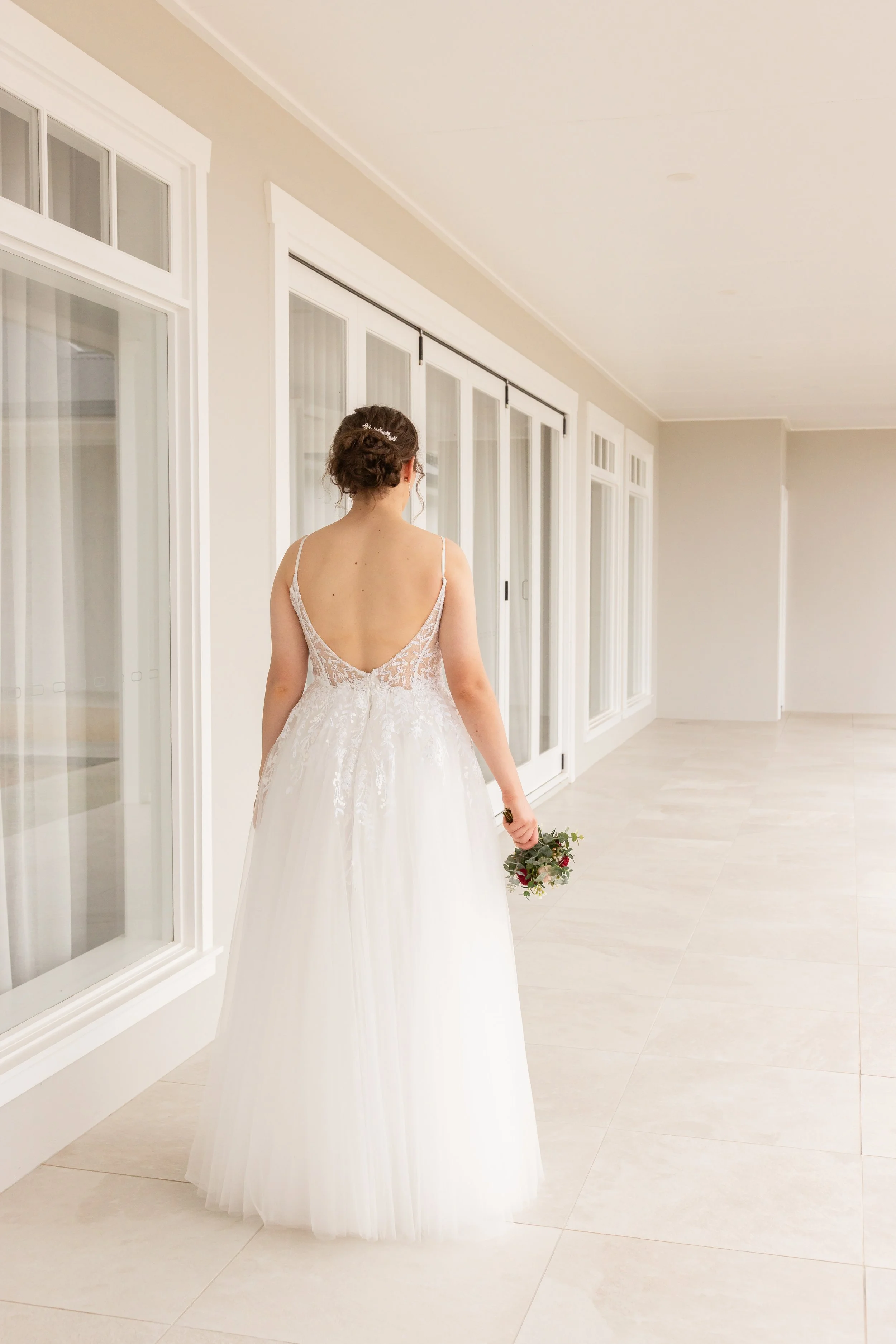 A woman in a white wedding dress holding a bouquet, standing in a bright, modern hallway with large windows.