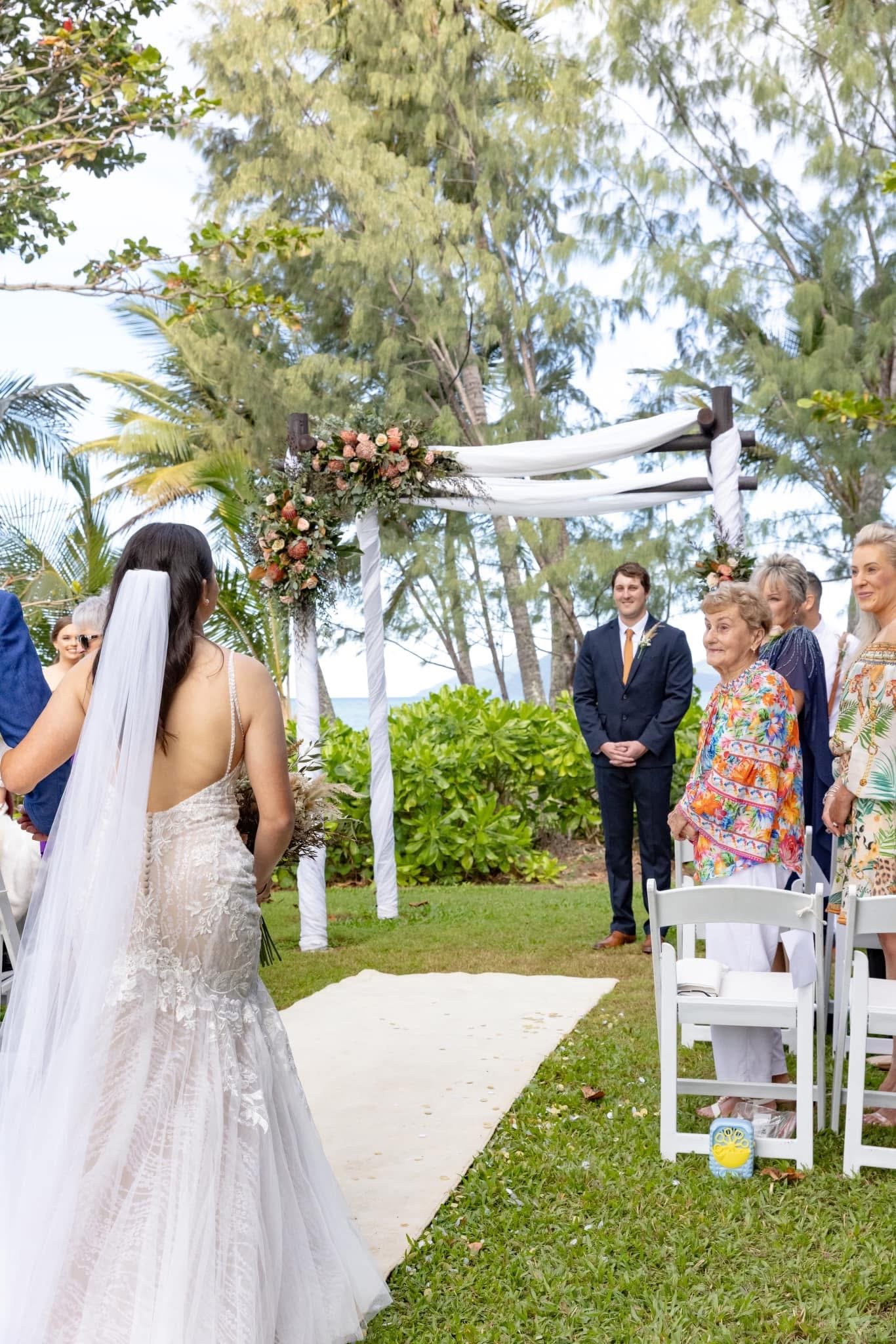 A wedding ceremony outdoors with a bride in a lace dress and guests standing under a decorated archway, surrounded by trees and greenery.