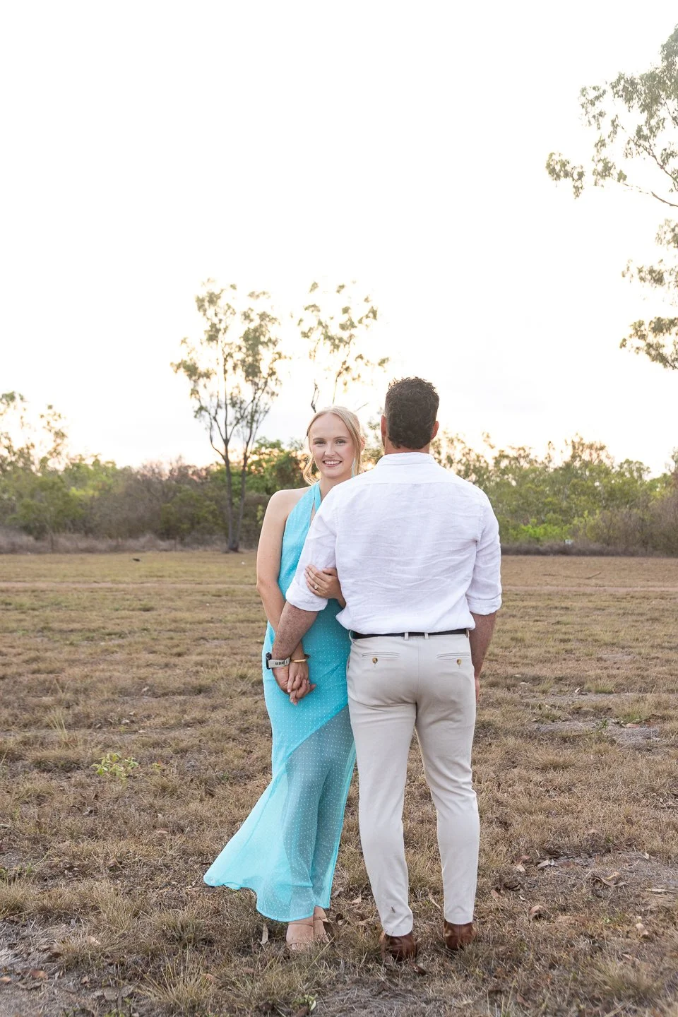 A woman in a blue dress holding hands with a man in white shirt and beige pants standing in an open field with trees in the background during sunset.