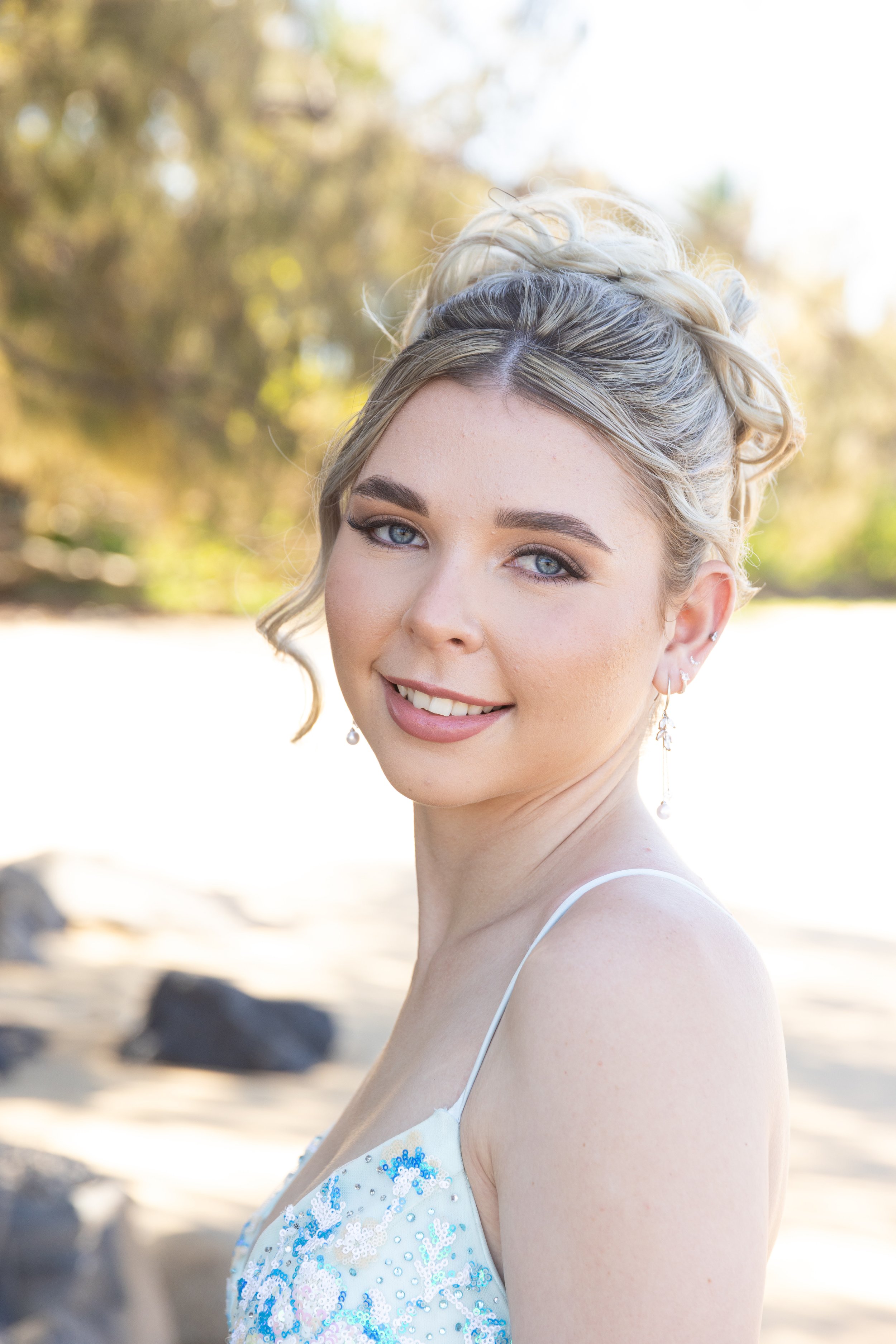 A young woman with blonde hair styled in an updo, wearing a white dress with blue sequins, earrings, and makeup, standing outdoors with blurred trees and rocks in the background.