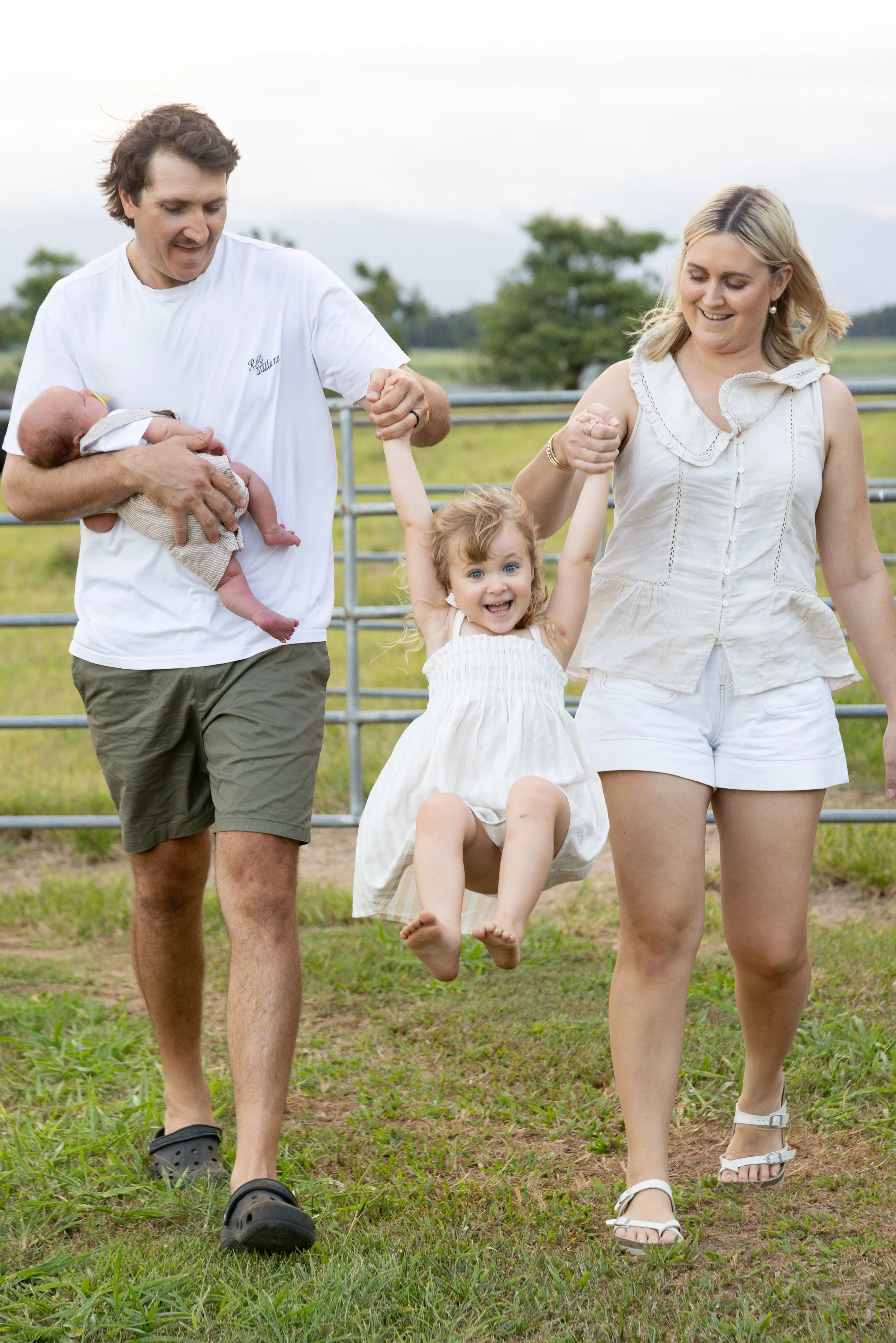 Family of four outdoors on farm, parents holding two children, one dad holding an infant and mom swinging a young girl with curly hair and a big smile.