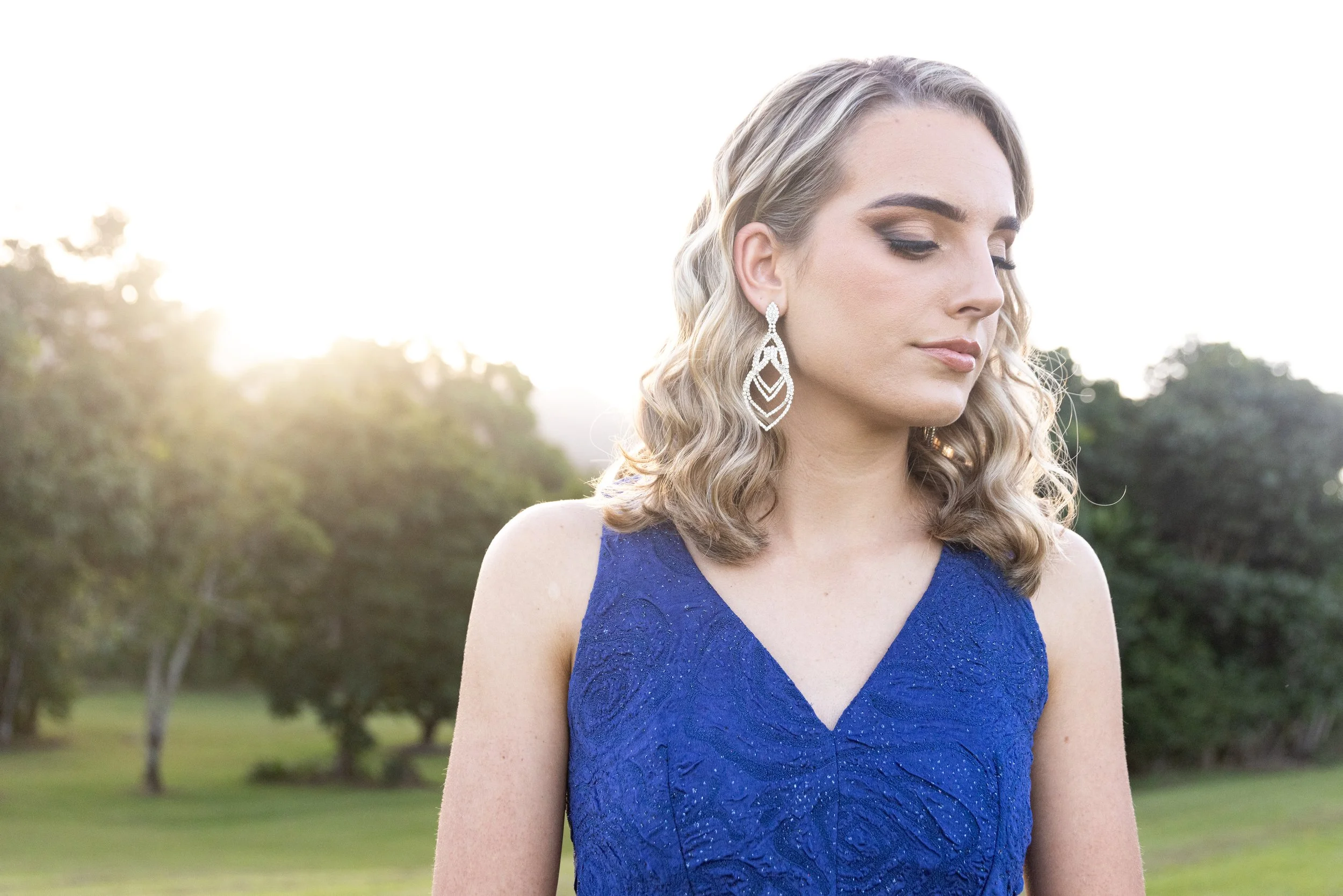 A young woman with wavy blonde hair wearing a royal blue dress and large, dangling diamond earrings stands outdoors in natural sunlight, with trees and a grassy area in the background.