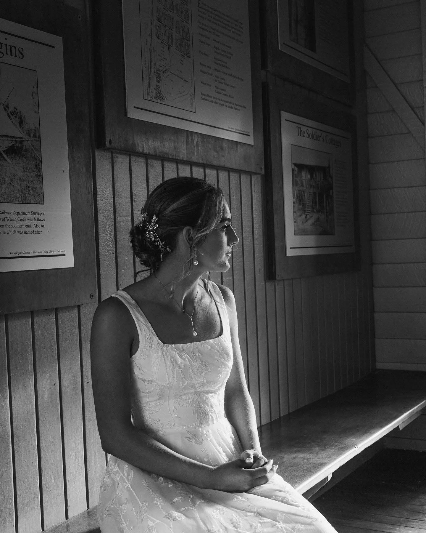 A woman in a white dress with embroidery sits on a wooden bench, looking to her left, in a room with wood-paneled walls and framed posters.