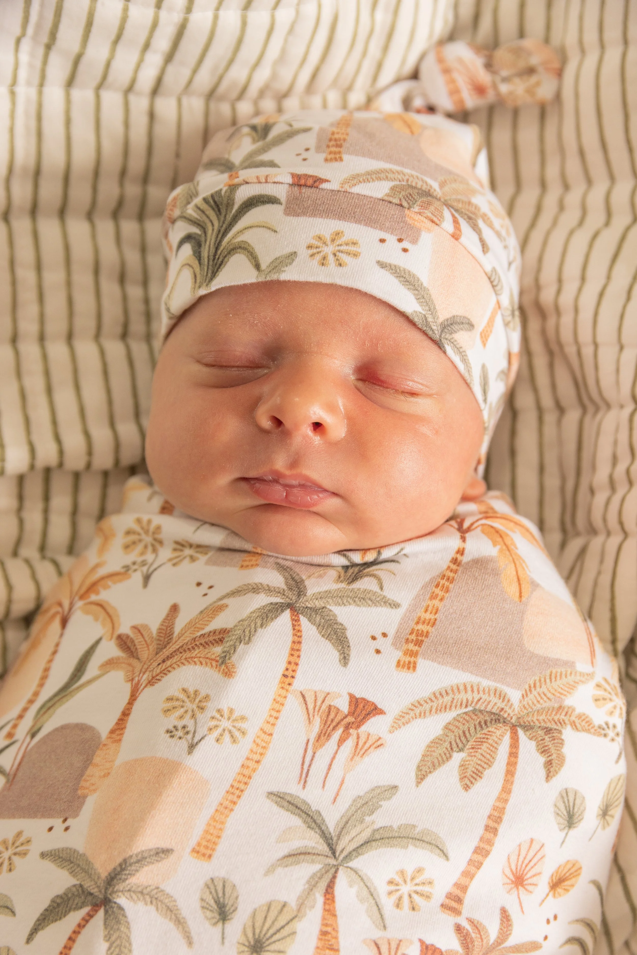 Close-up of a newborn baby sleeping peacefully, wrapped in a blanket with a tropical palm tree pattern, wearing a matching hat, lying on a striped blanket.