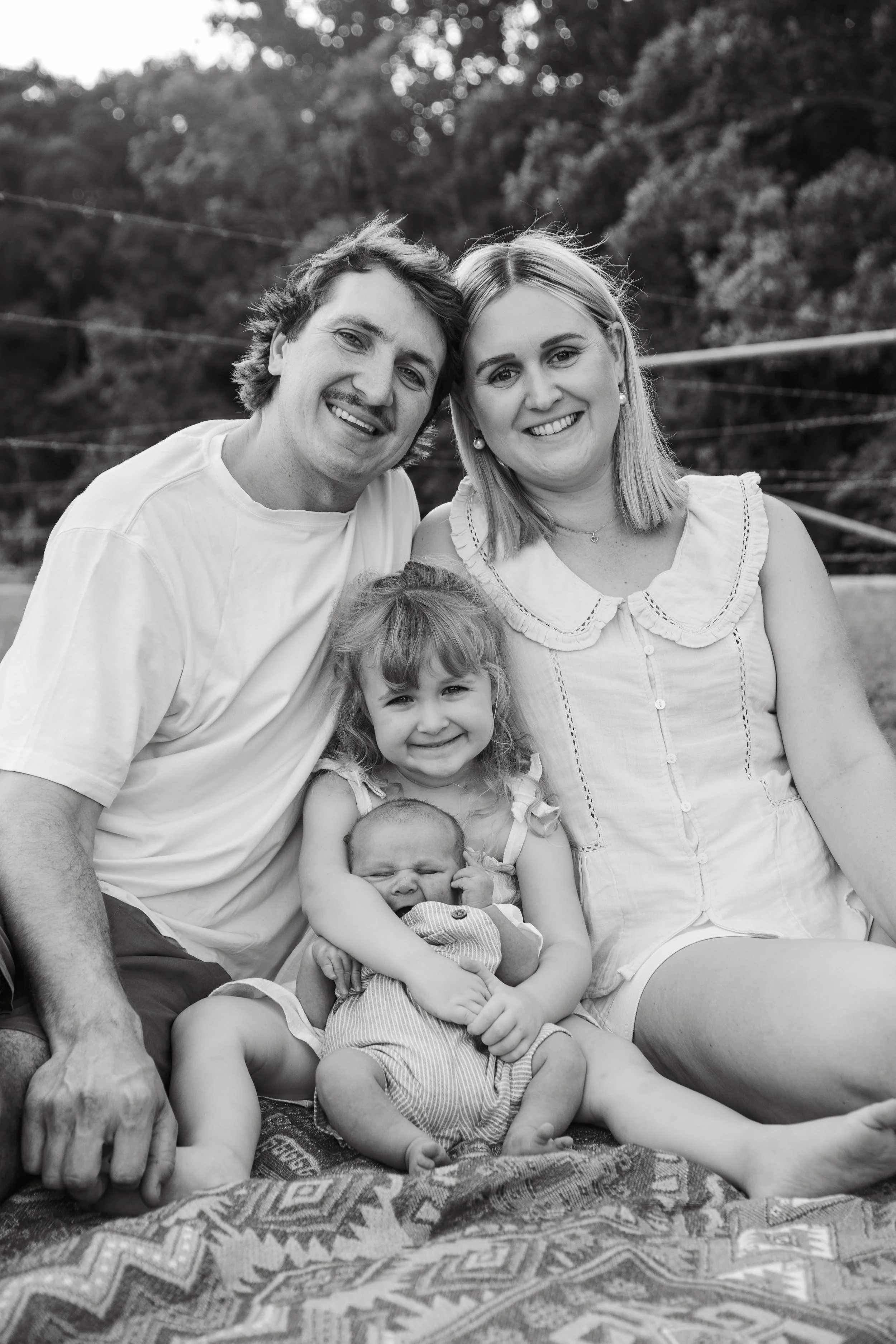 Black and white photo of a smiling family outdoors, including a man, woman, young girl, and baby, sitting on a blanket with trees in the background.