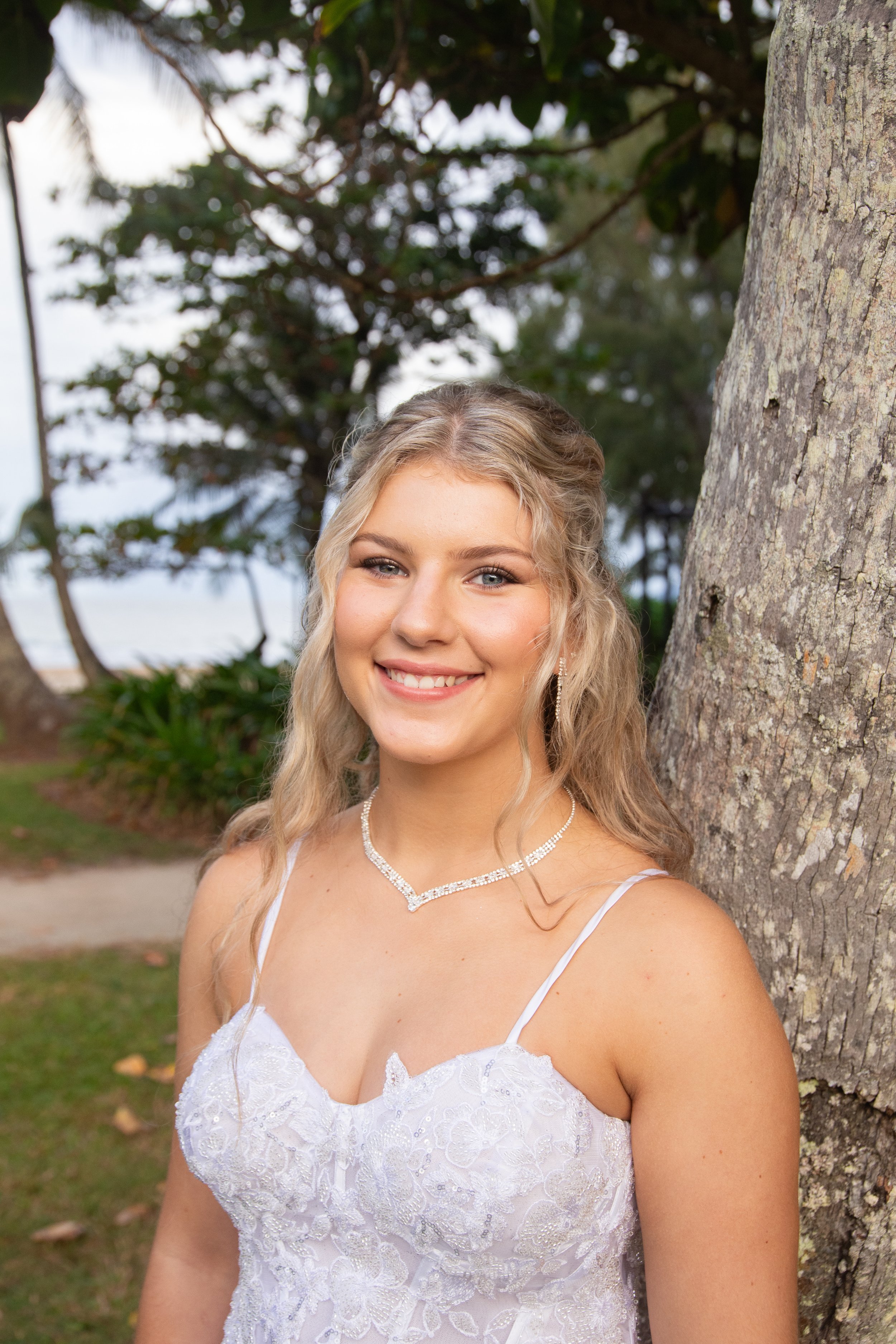 A young woman in a white lace dress with thin straps standing outdoors next to a tree, smiling at the camera. She is wearing pearl jewelry and has long, wavy blonde hair.