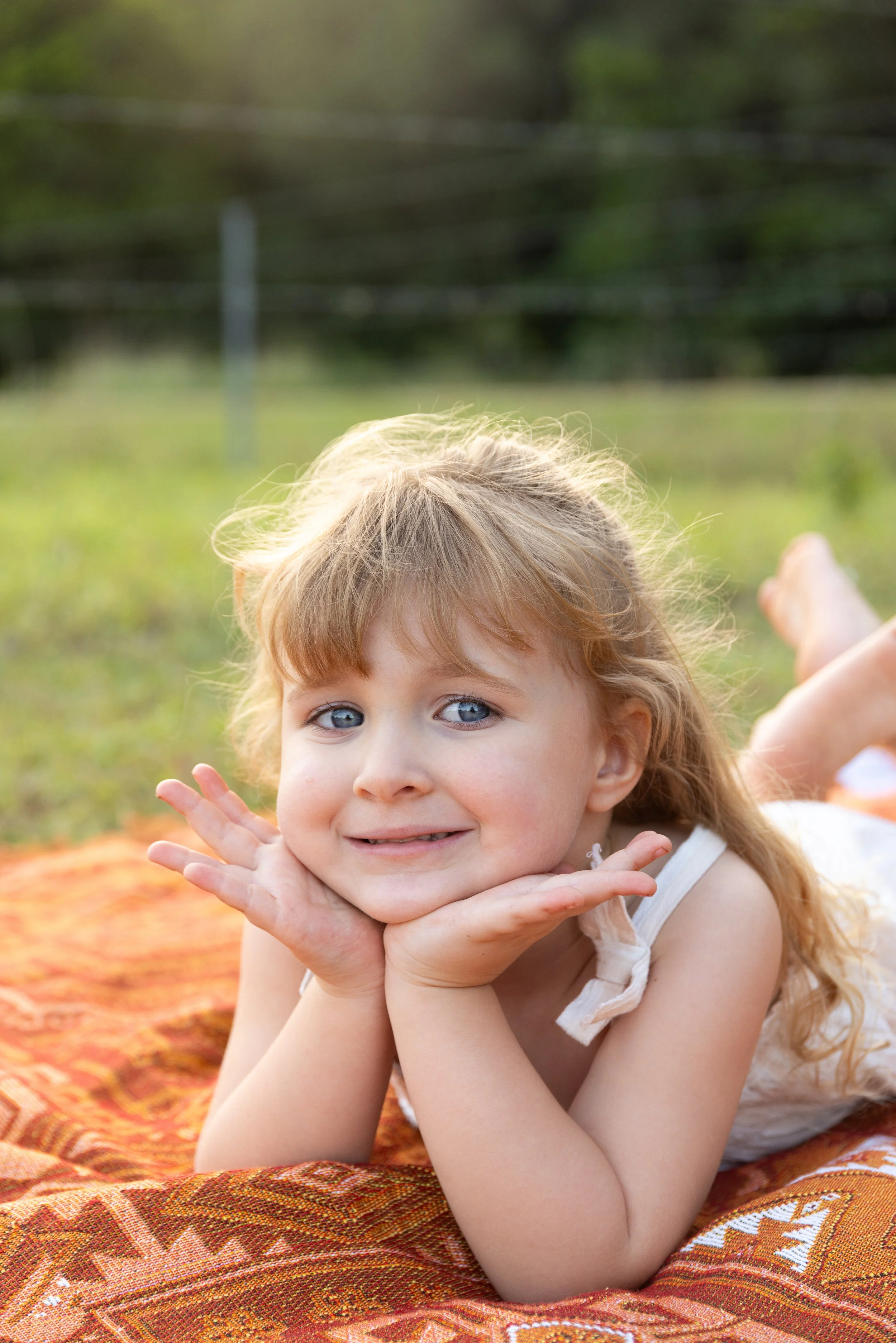 A young girl with blue eyes and long blonde hair lying on a patterned blanket outdoors with grass and trees in the background.