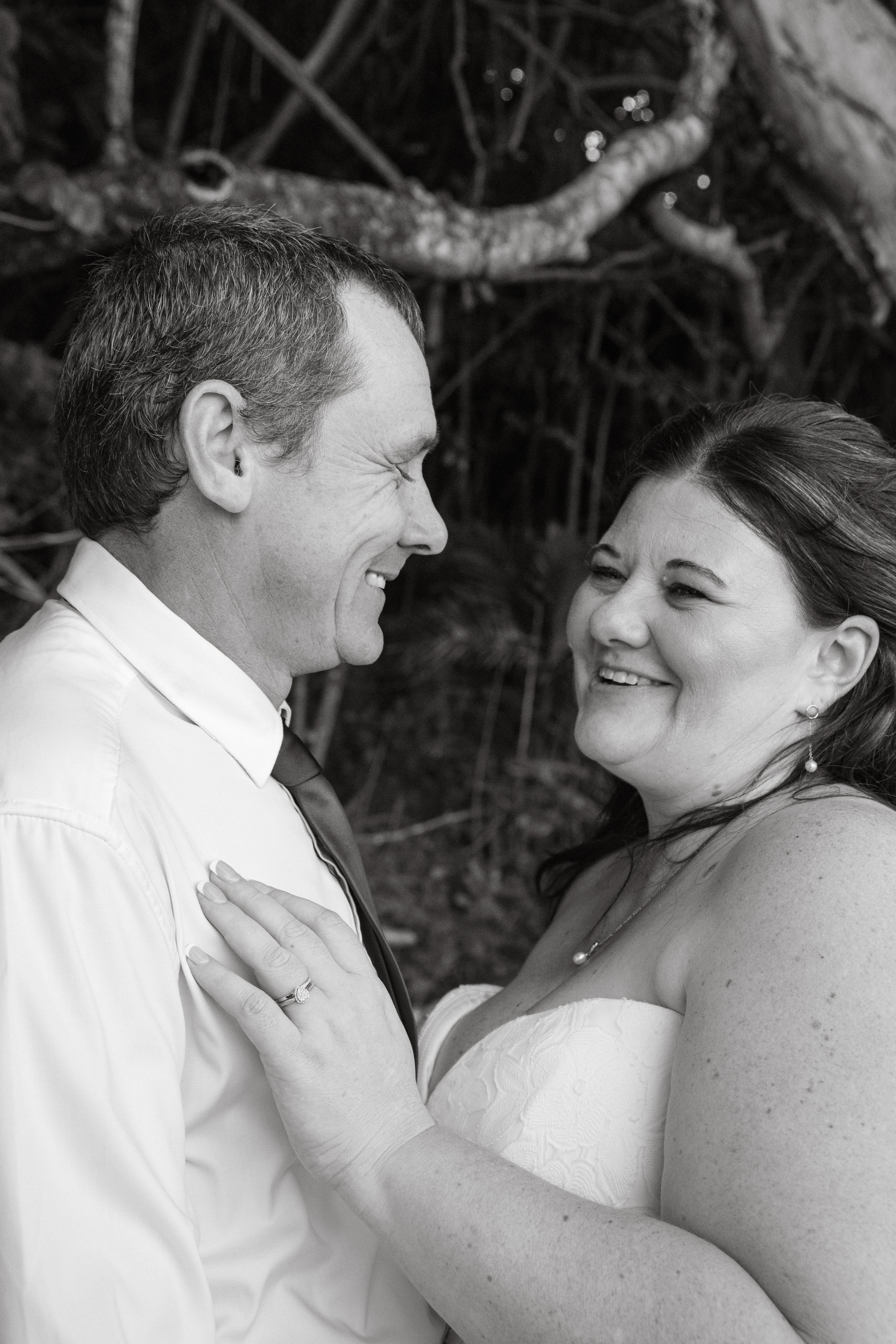 A happy couple on their wedding day, smiling and looking at each other, outdoors with trees in the background. The woman wears a strapless dress and the man is dressed in a shirt and tie.
