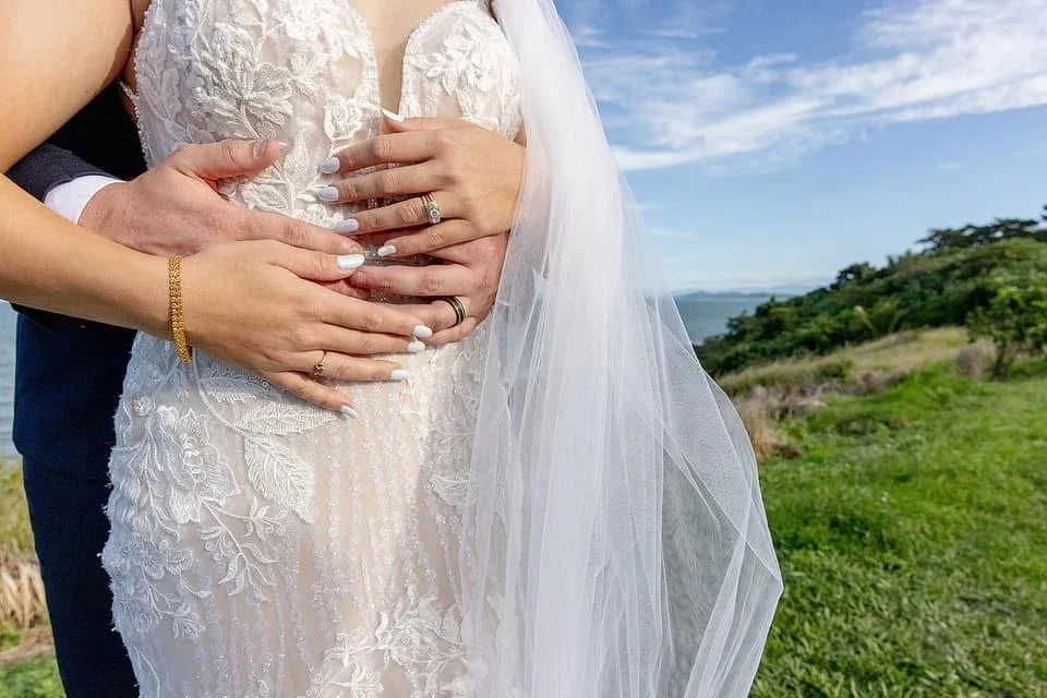 Close-up of a bride and groom holding her pregnant belly at an outdoor wedding, with a scenic landscape of greenery and water in the background.