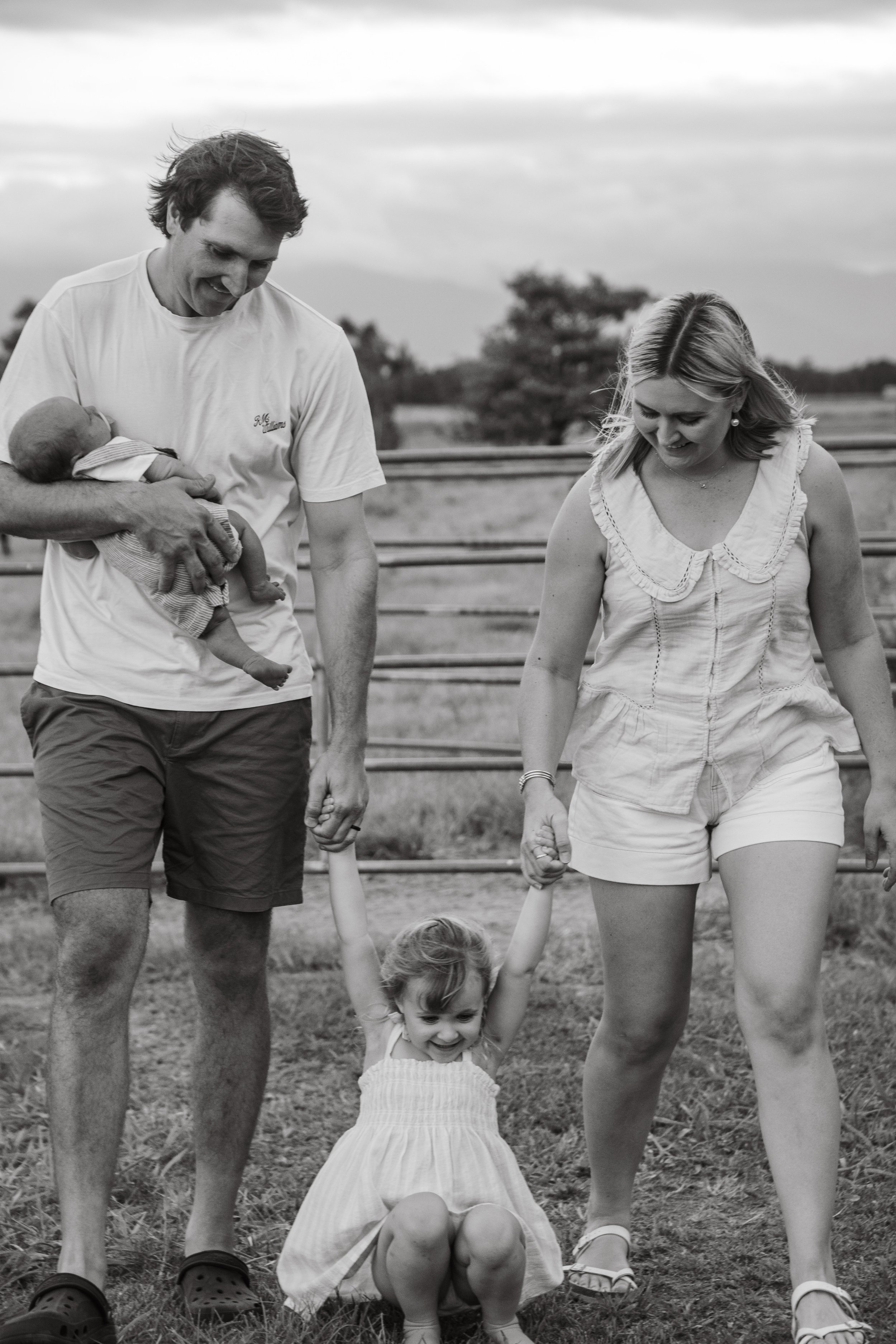 A black-and-white photo of a young girl smiling and sitting on the grass, holding hands with a man and a woman, possibly her parents, outdoors near a metal fence with trees and cloudy sky in the background.