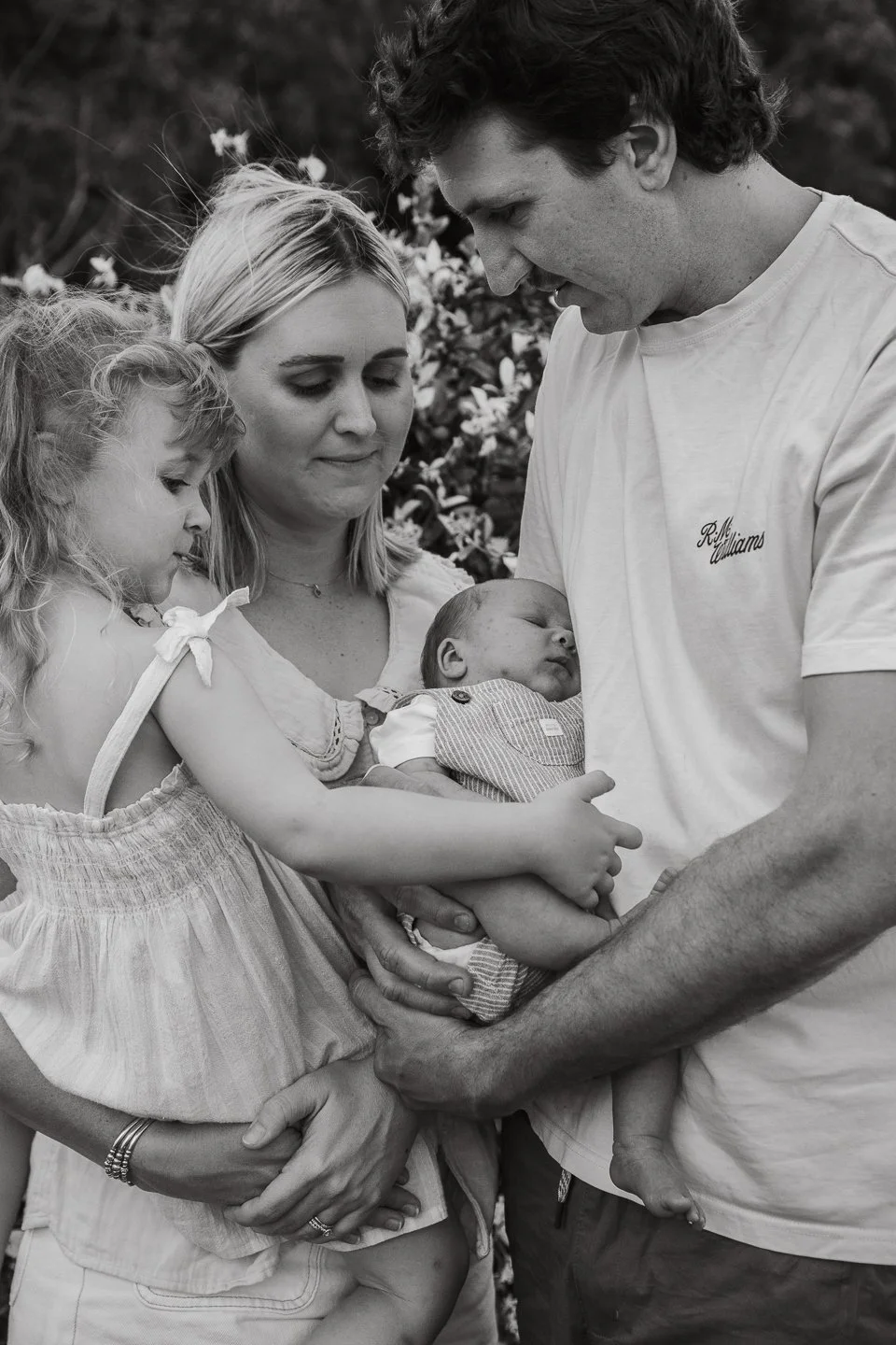 A black and white photo of a family holding a newborn baby with two young girls, one of them sleeping on her mother, watching the baby as the father cradles the infant.