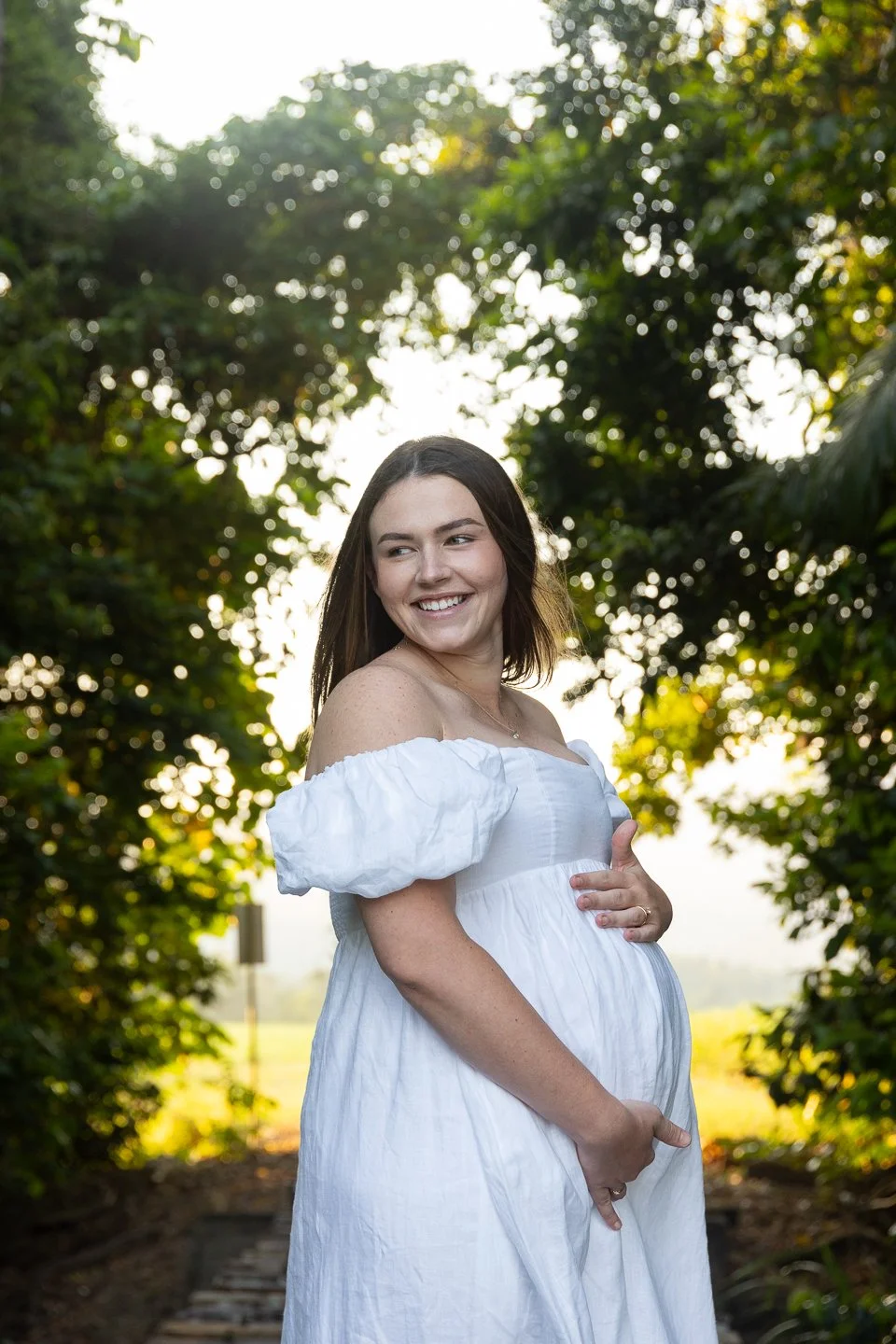 A pregnant woman with long brown hair, smiling, wearing a white off-shoulder dress, stands outdoors in a wooded area during the daytime with sunlight filtering through the leaves.