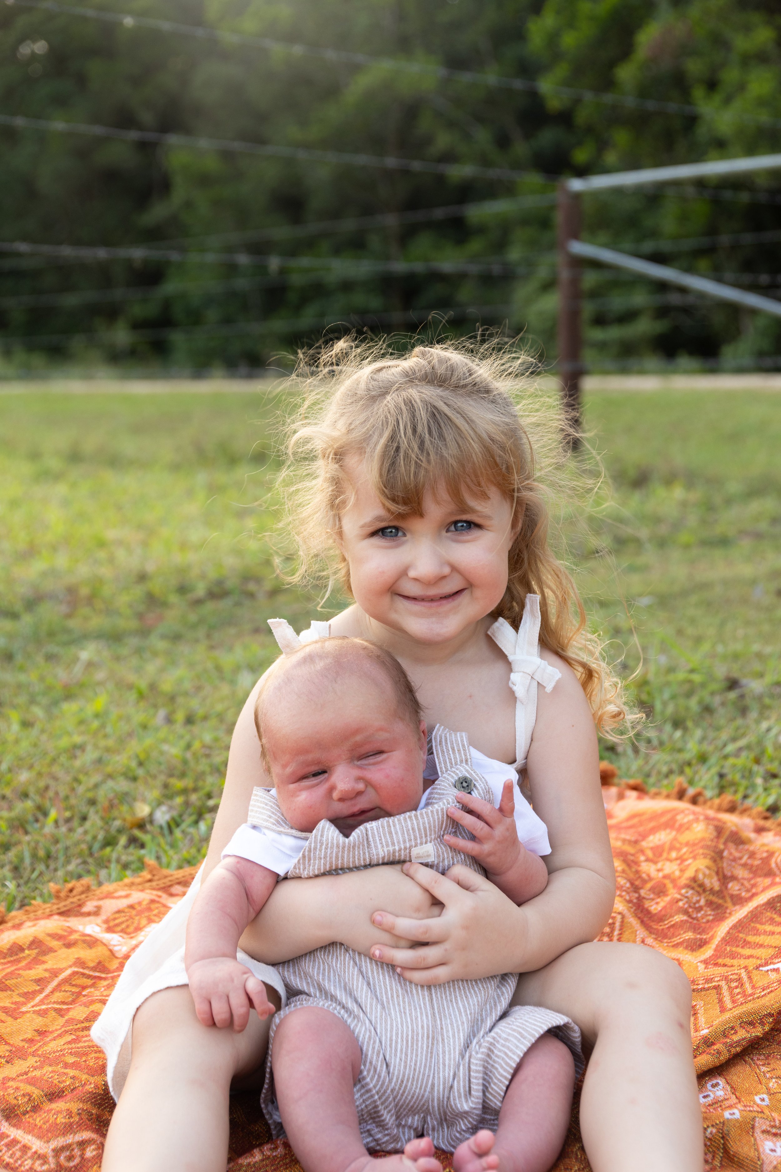 A young girl with curly blonde hair and blue eyes smiling while holding a newborn baby with light brown hair. They are sitting on an orange blanket outdoors on green grass with trees and power lines in the background.