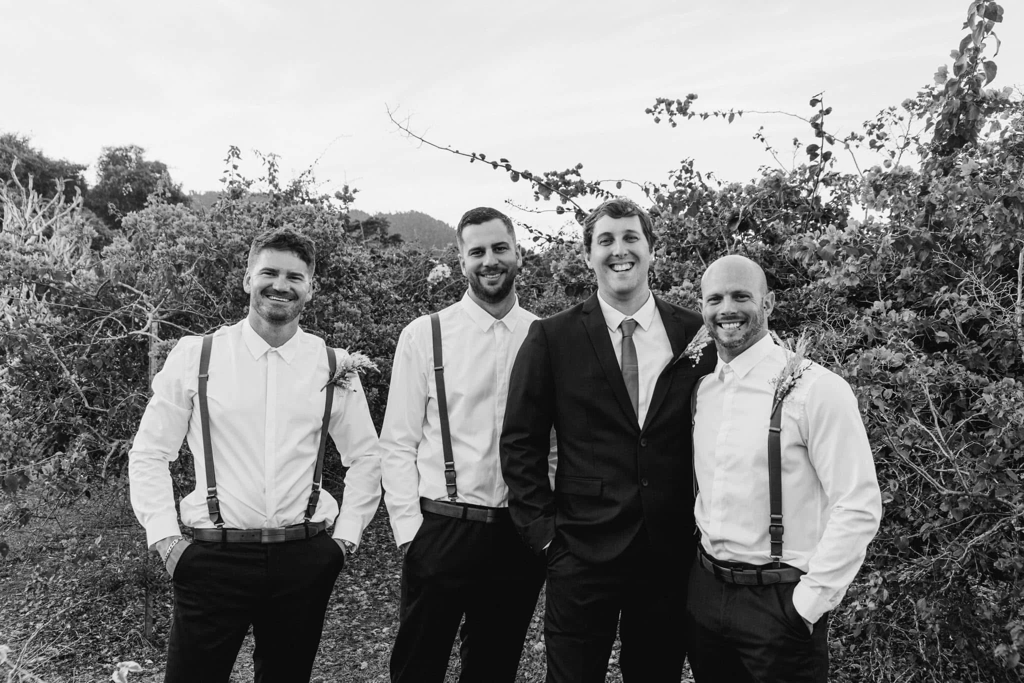 Black and white photo of four men dressed in formal attire, standing outdoors with trees and mountains in the background. The man in the center is wearing a suit and tie, while the others are in white shirts with suspenders and boutonnières, smiling 