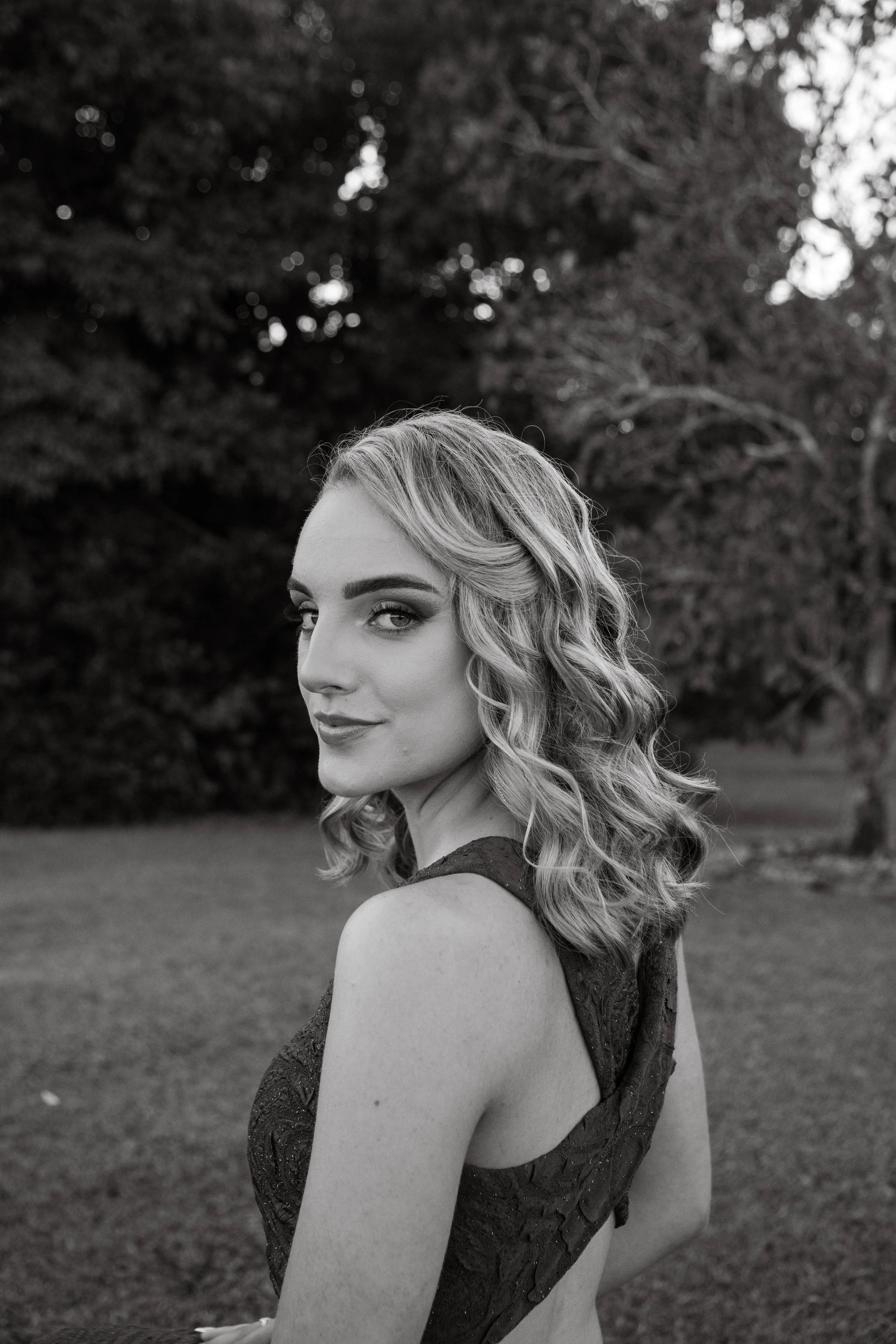 A black-and-white portrait of a woman with wavy shoulder-length hair, wearing a sleeveless dress, standing outdoors with trees and grass in the background.