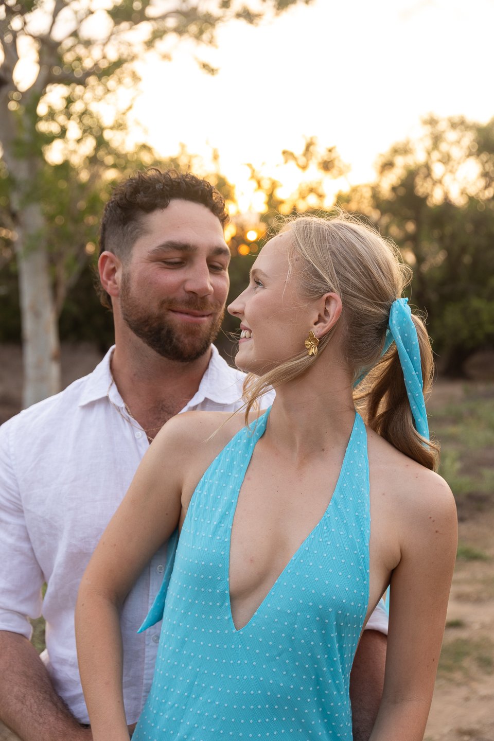 A young couple smiling at each other outdoors during sunset, with trees in the background. The woman wears a blue polka dot halter top and a matching headband, and the man wears a white shirt.