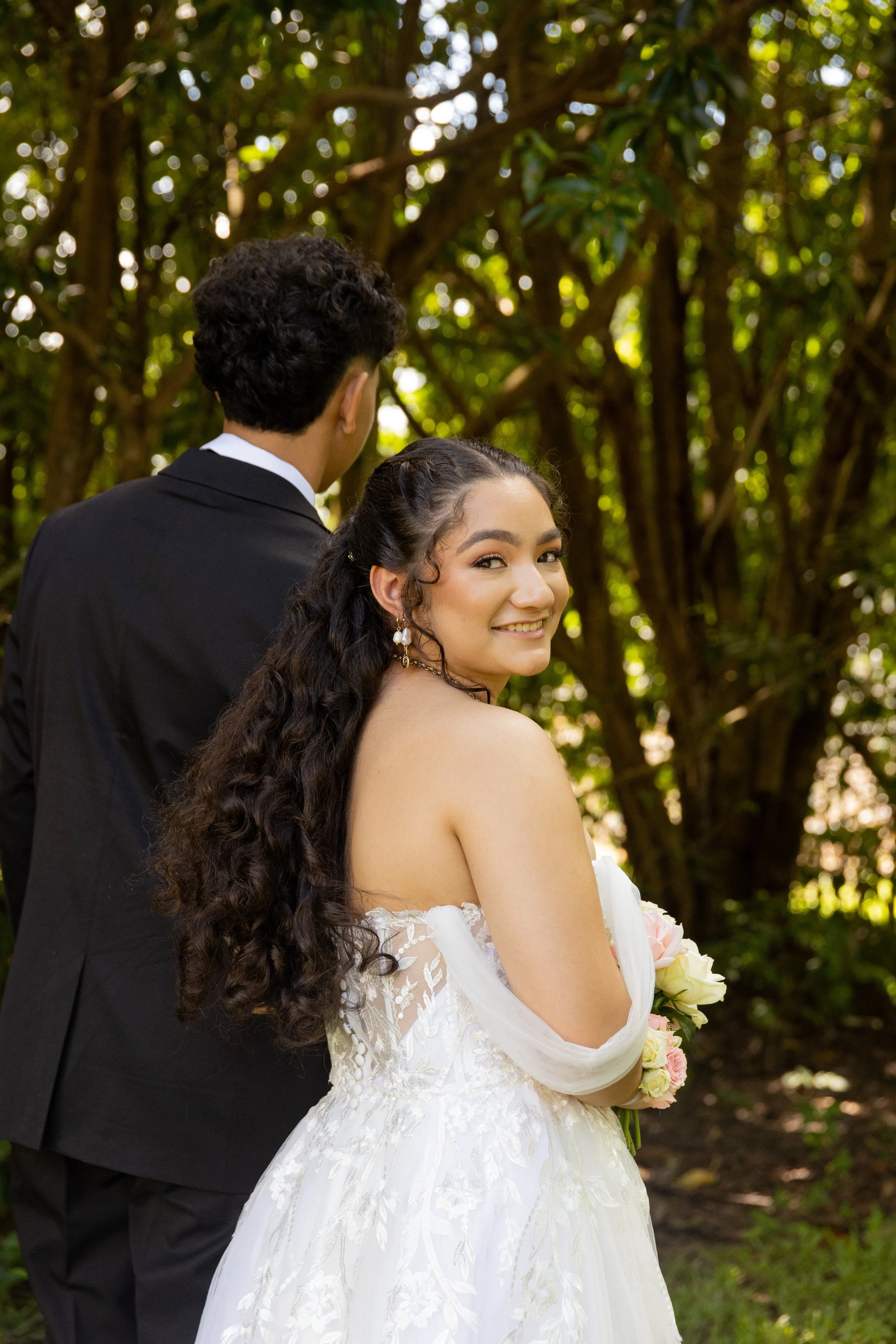 A bride with long curly hair in a white wedding dress holding a bouquet, smiling and looking over her shoulder, standing outdoors with trees in the background. A groom in a black suit is beside her, facing away from the camera.