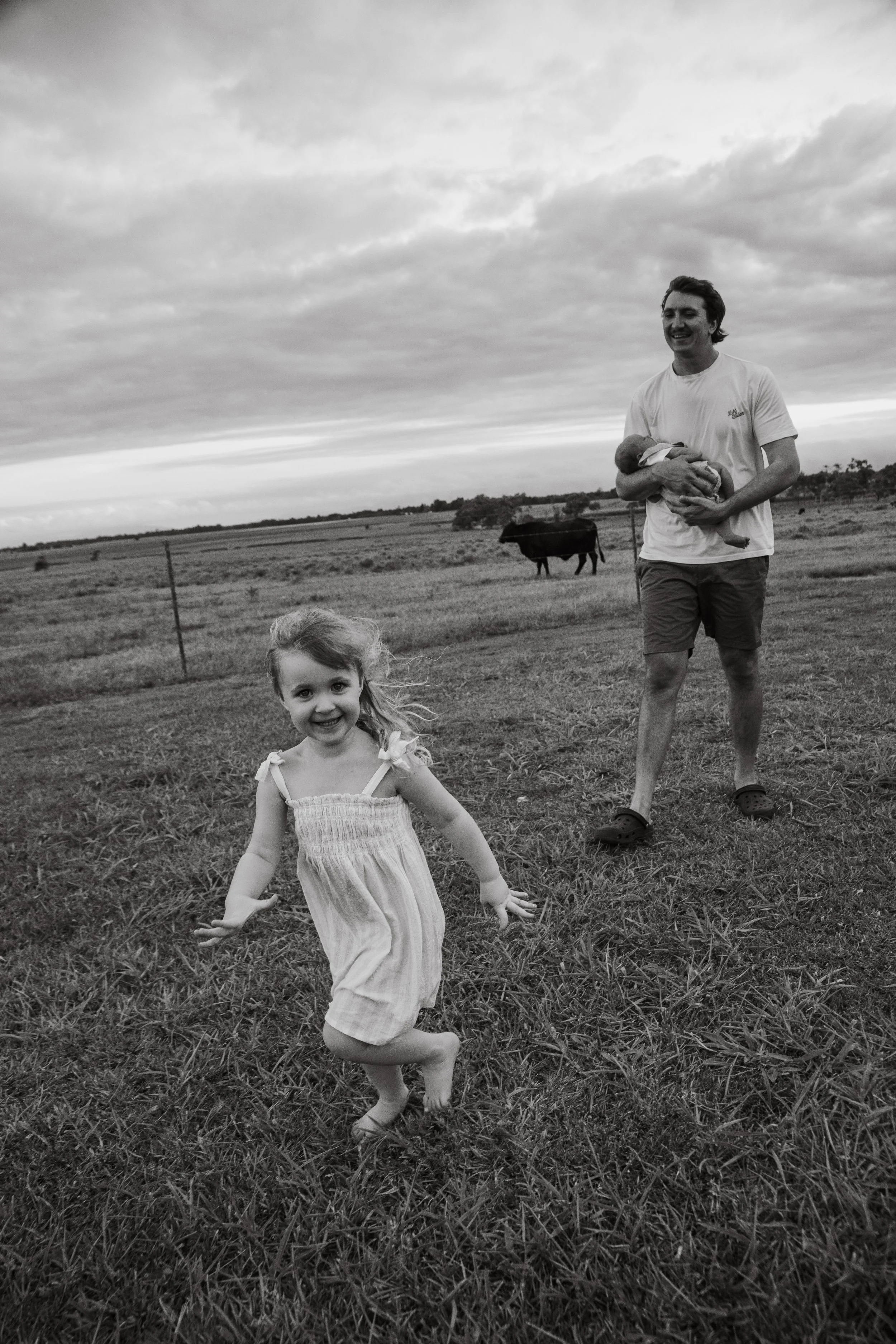 A black and white photograph of a man holding a newborn baby and a young girl running on a grassy field with cows in the background, under a cloudy sky.
