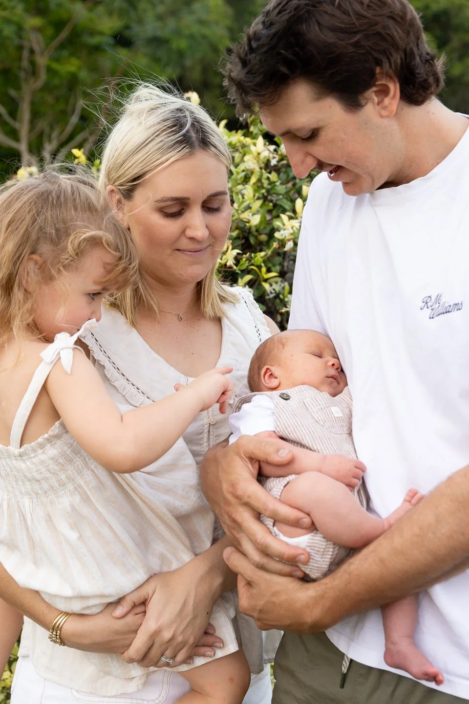 A family of four, including a mother, father, young girl, and infant, standing outdoors in a garden with green foliage, sharing a tender moment as they look at and hold the sleeping baby.