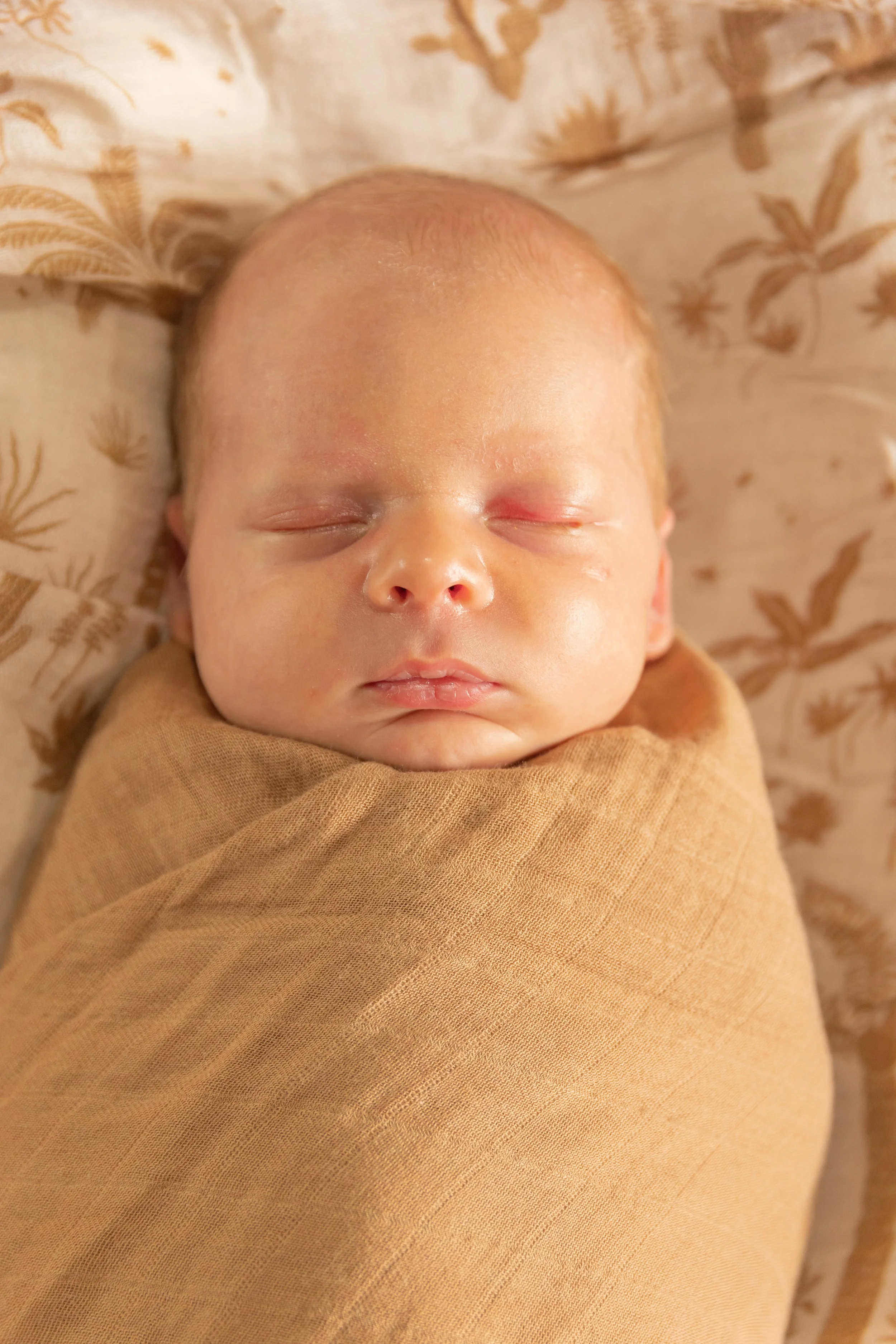 A sleeping newborn baby wrapped in a light brown cloth, lying on a patterned beige and gold blanket.