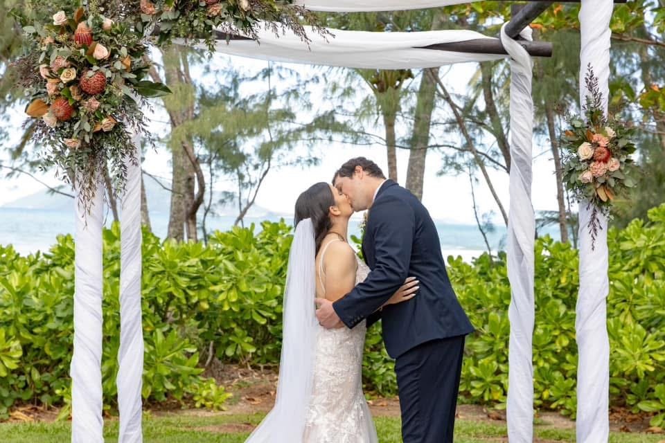 A bride and groom share a kiss under a wedding arch decorated with flowers and greenery, outdoors near a body of water with trees in the background.