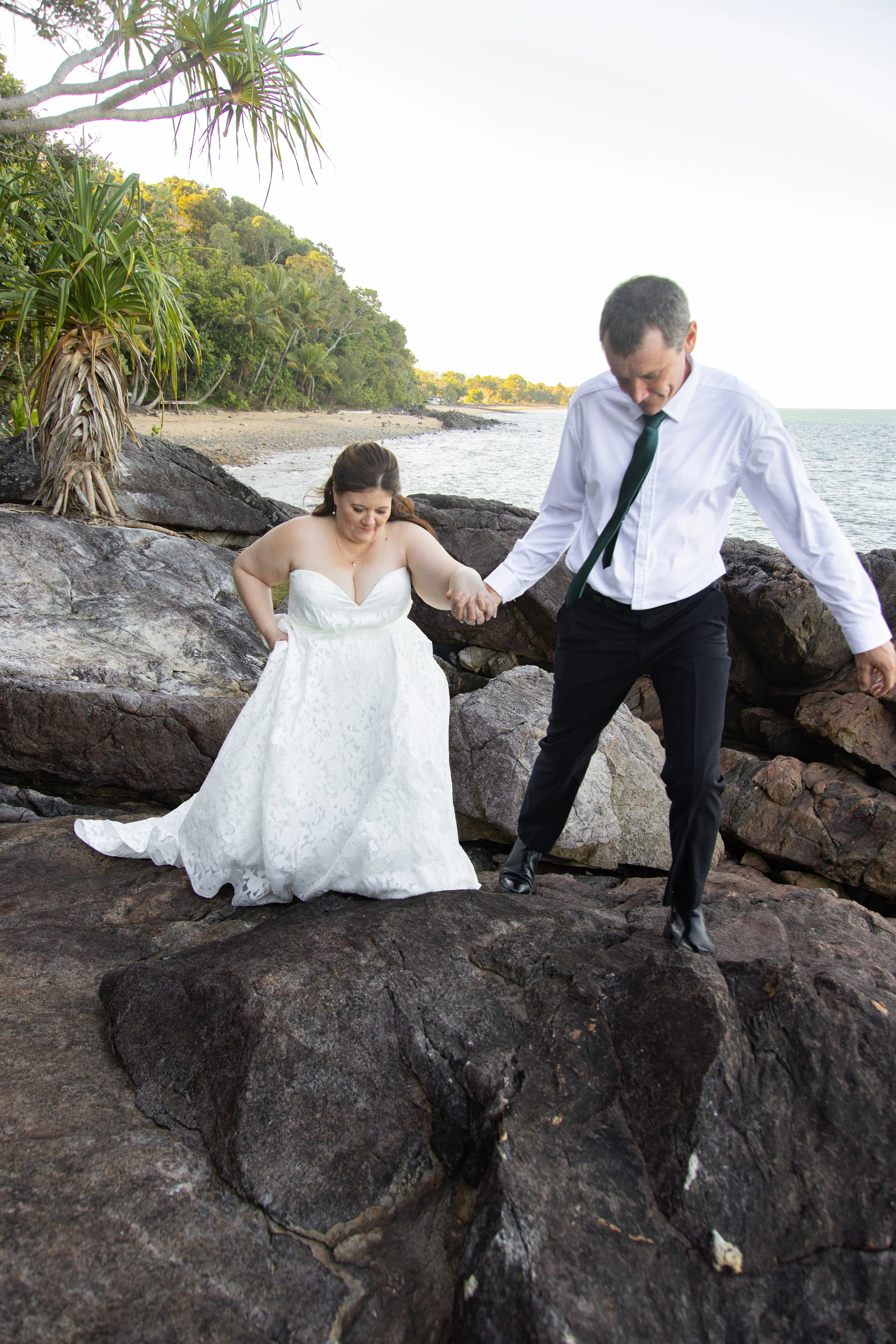 A bride in a white gown and a groom in a white shirt and black trousers walk on rocks by the ocean, holding hands.