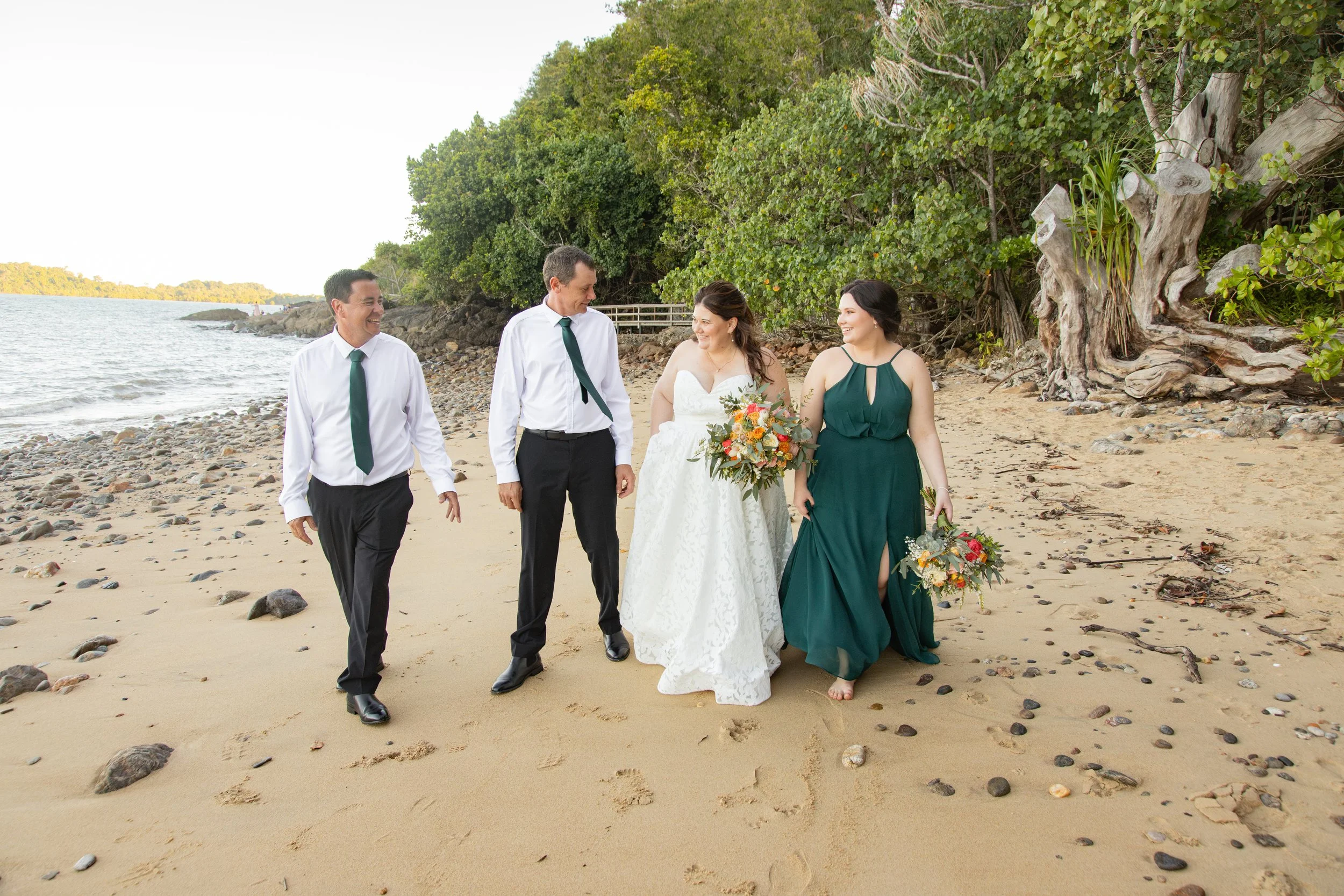 Group of five people dressed in wedding attire walking along a sandy beach with rocks, trees, and water in the background.