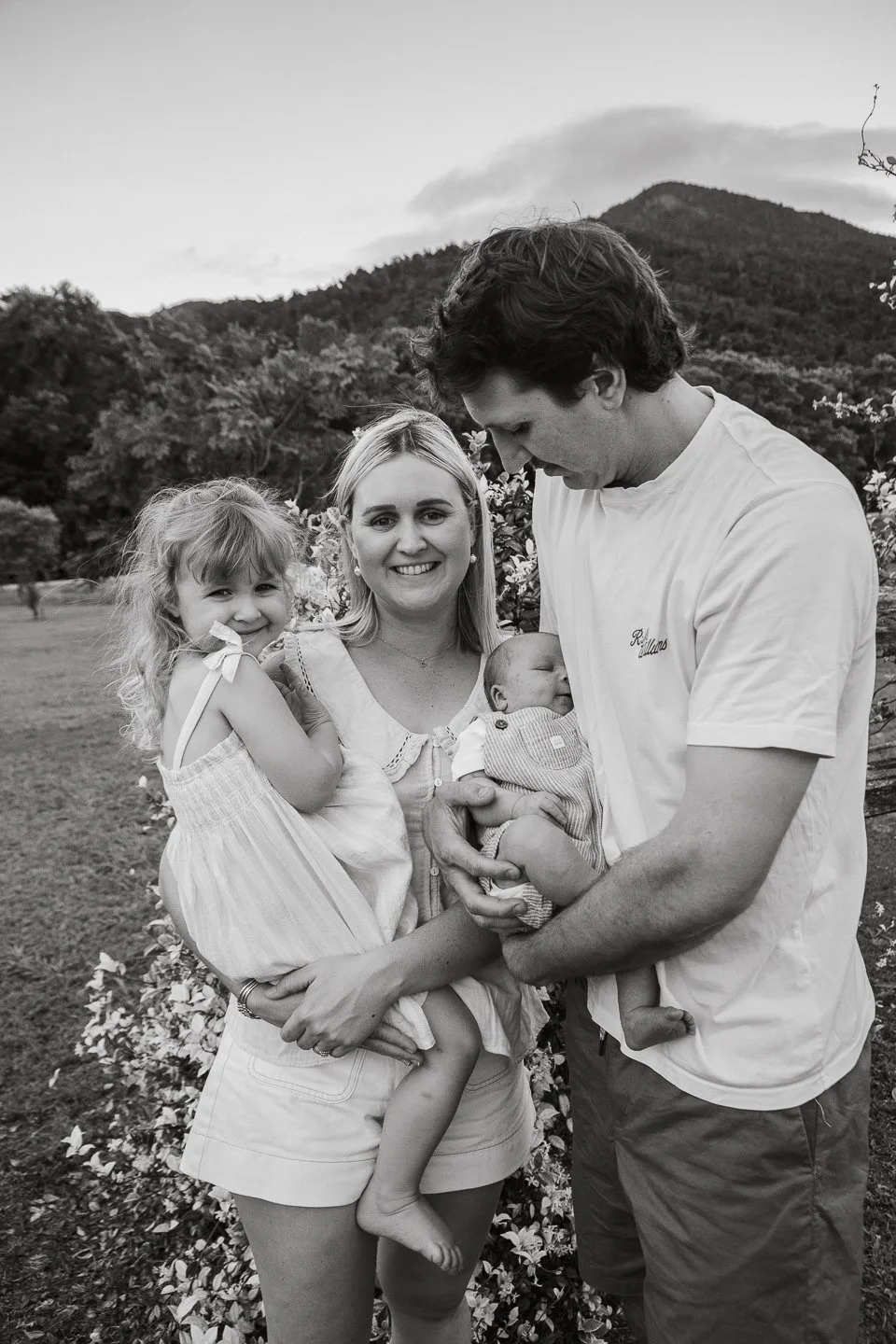 A family of four poses outdoors with mountains in the background. The woman holds a young girl, and a man holds a newborn baby. All are smiling.