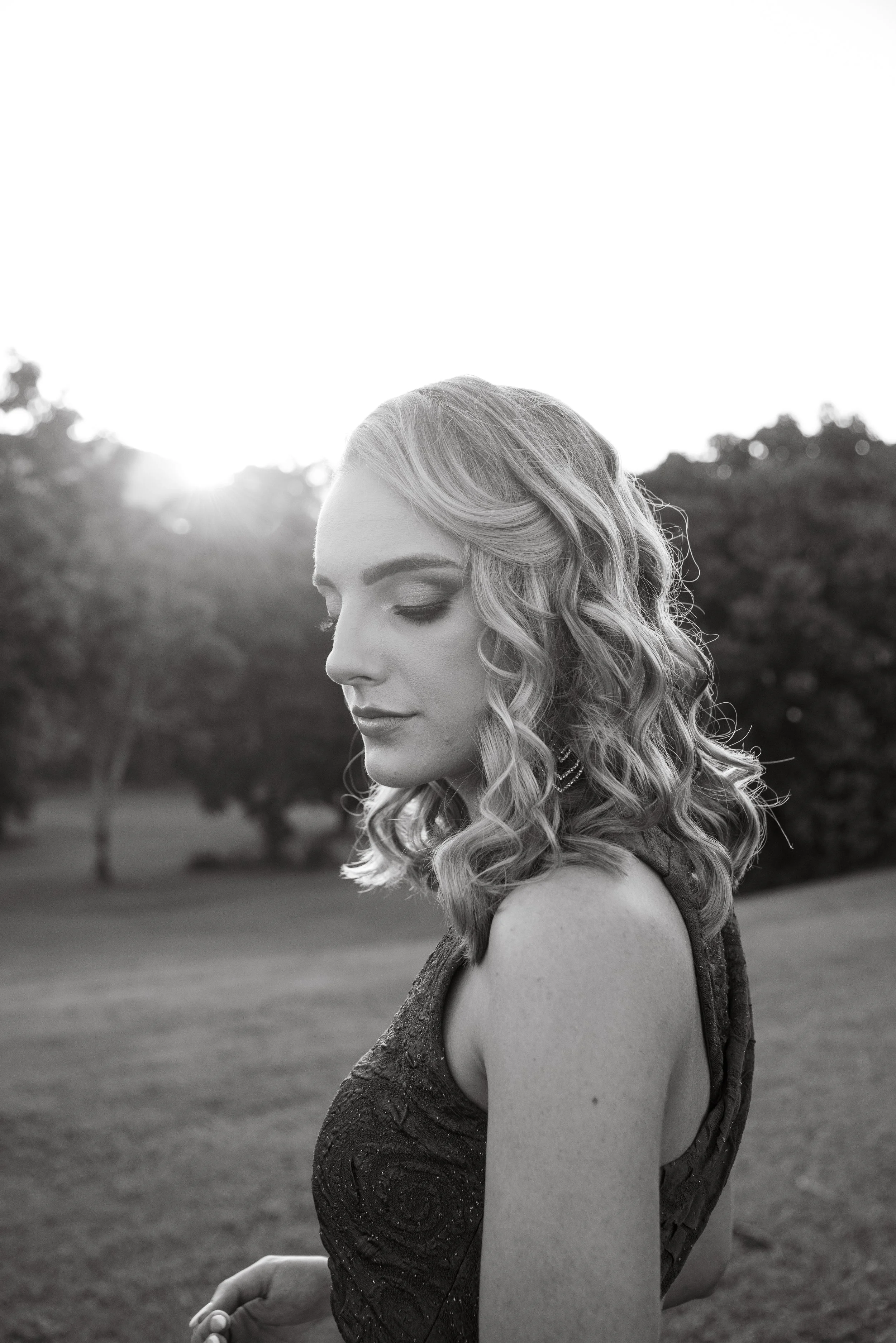 A woman with curly hair and closed eyes stands outdoors at sunset, wearing a sleeveless dress.