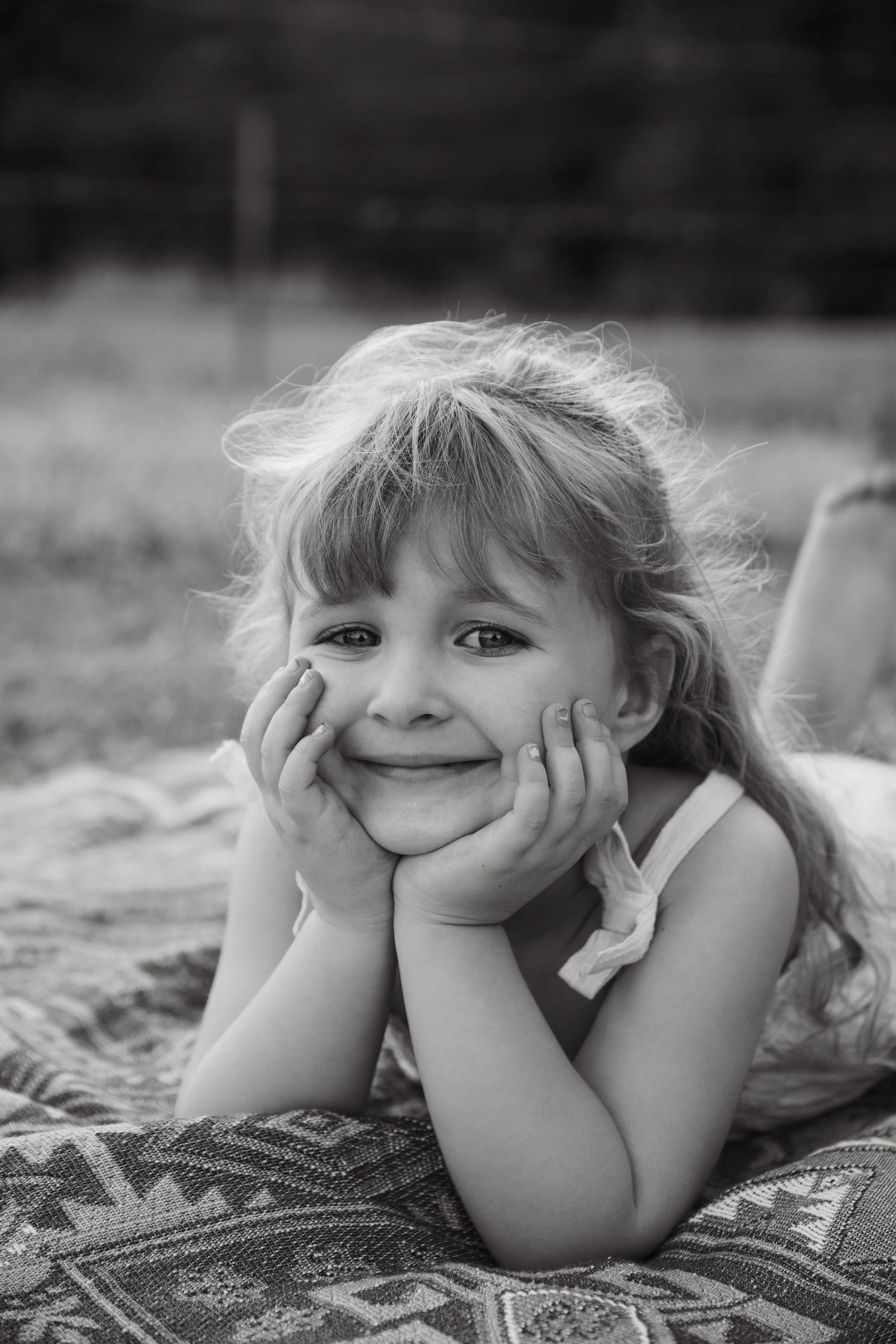 A young girl with curly hair resting her chin on her hands and smiling at the camera while lying on a patterned blanket outdoors.