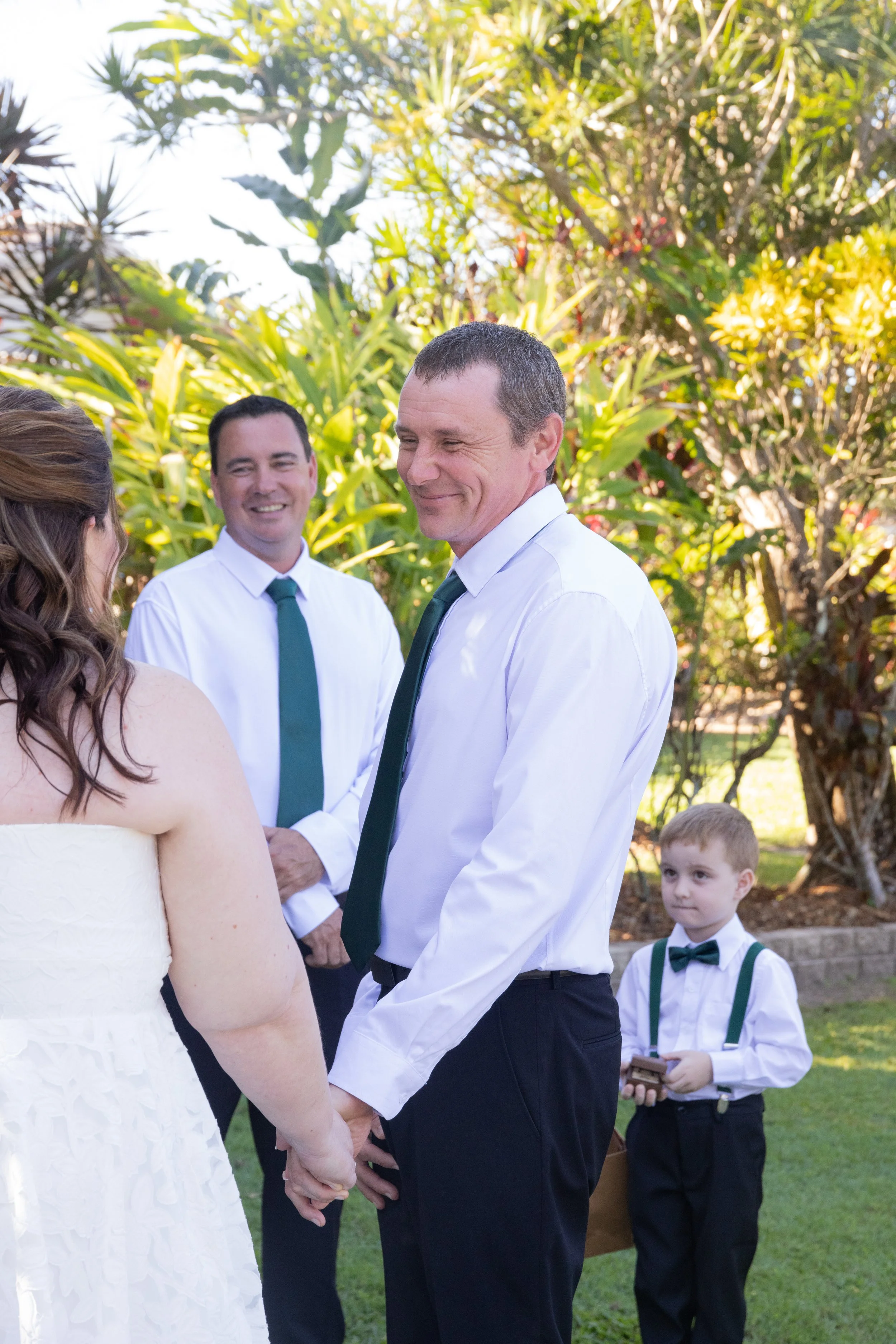 A wedding ceremony taking place outdoors with a bride, groom, and two men officiating or witnessing, all dressed in formal attire, with lush green foliage in the background.