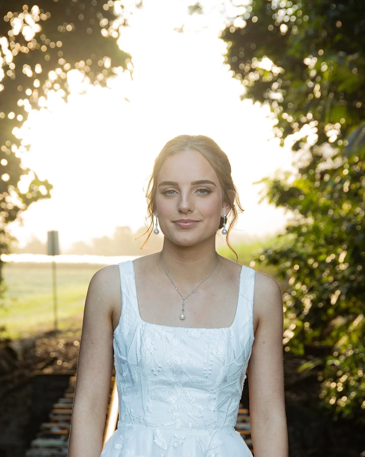 A young woman with light hair and blue eyes wearing a white dress and jewelry, standing outdoors on a sunny day with trees in the background.