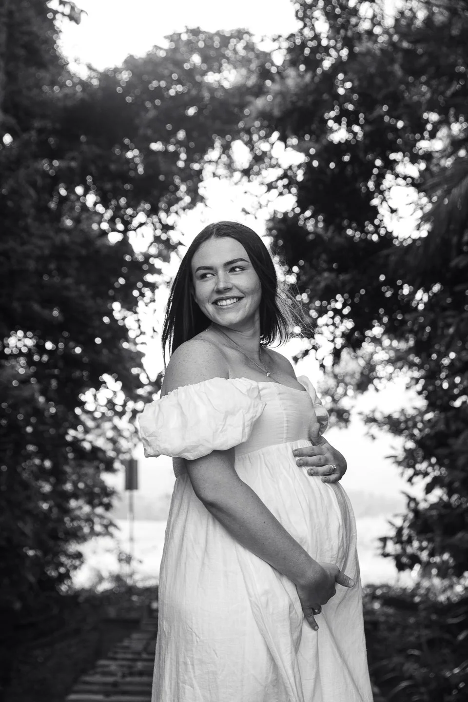 Black and white photo of a smiling pregnant woman standing outdoors, wearing a white off-shoulder dress with puffy sleeves and holding her belly with one hand, with trees and a sky background.