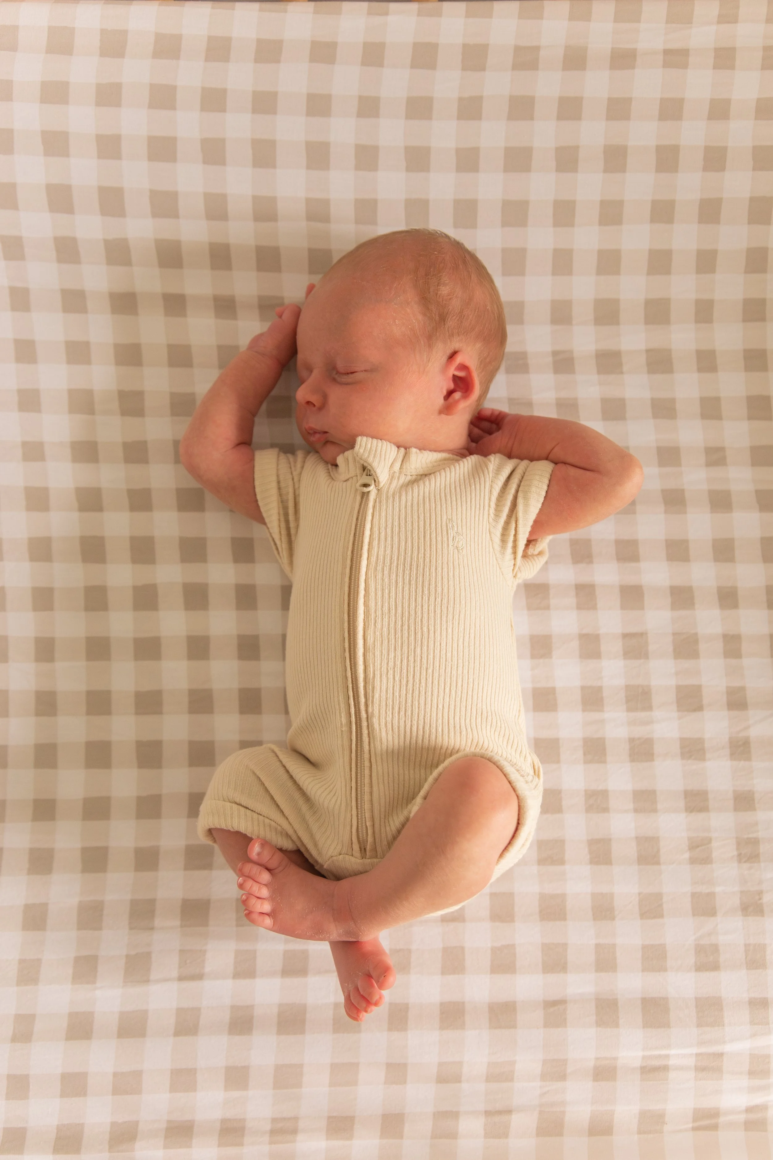 A newborn baby sleeping peacefully on a checkered beige and white blanket, dressed in a beige zip-up onesie.