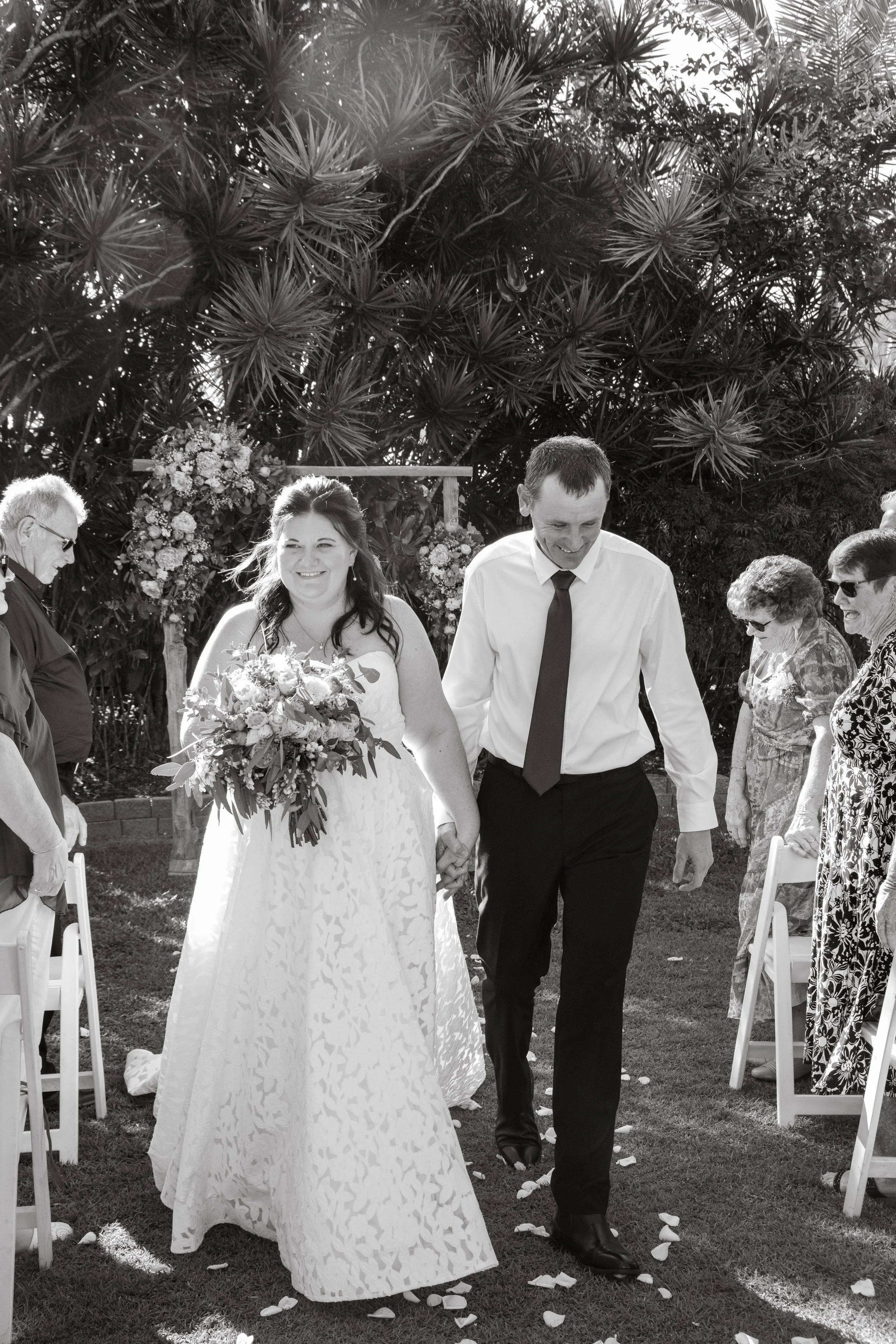 A couple holding hands and walking down the aisle during their outdoor wedding ceremony, surrounded by guests and floral decorations.