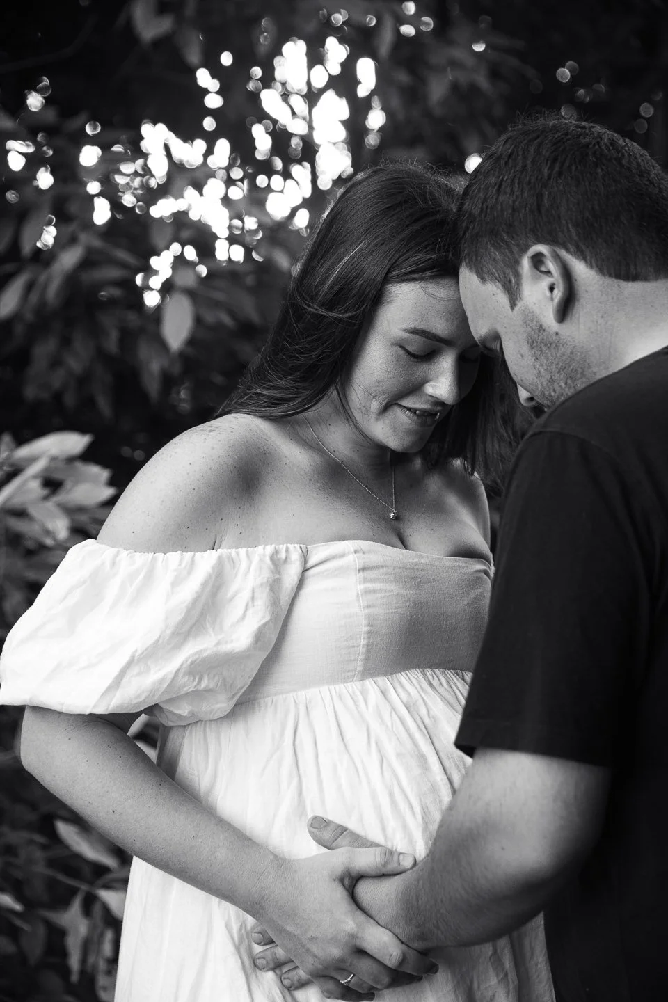 A black and white photograph of a pregnant woman and a man holding her hands and gently touching her belly, with their foreheads touching and eyes closed, outdoors with blurred foliage background.