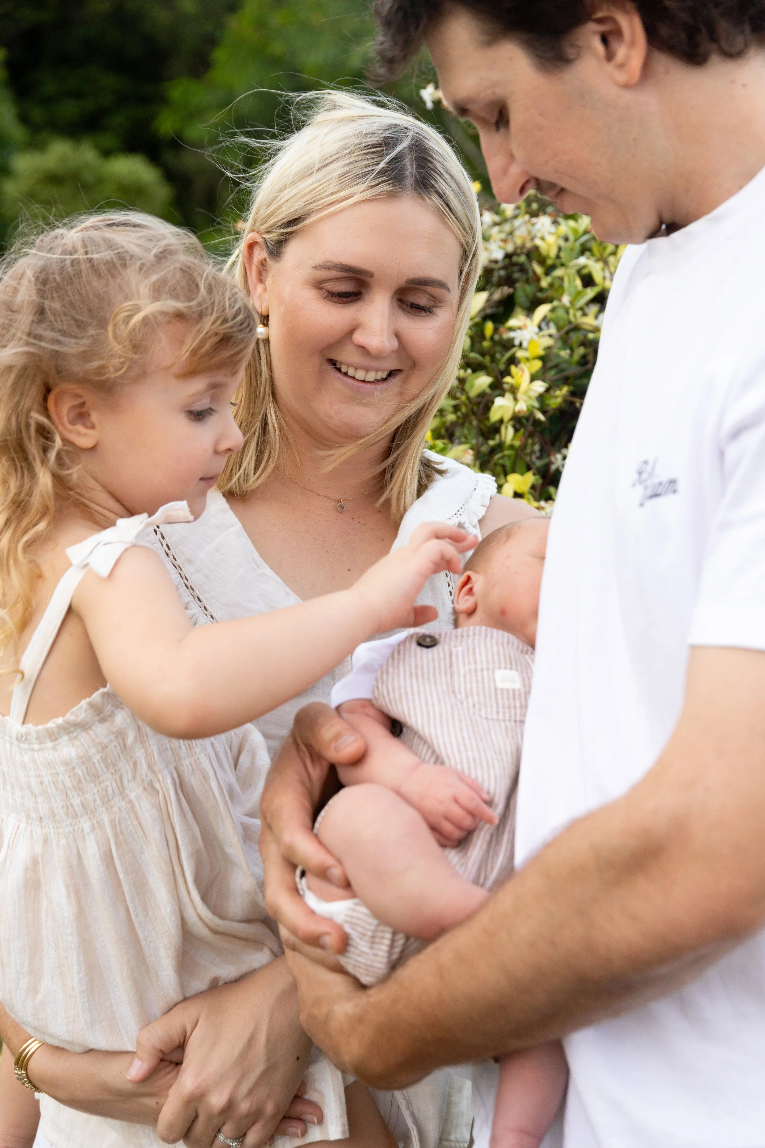 A family of four, including a mother with blonde hair, a father with dark hair, a young girl with blonde hair, and a newborn baby, are outdoors in a garden. The mother and father are holding the baby while the young girl touches the baby's head gentl