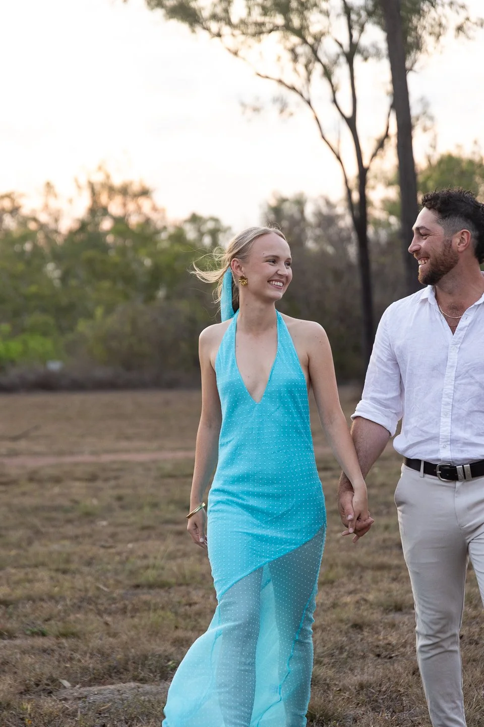 A smiling woman in a light blue dress holding hands with a man in a white shirt outdoors at sunset.