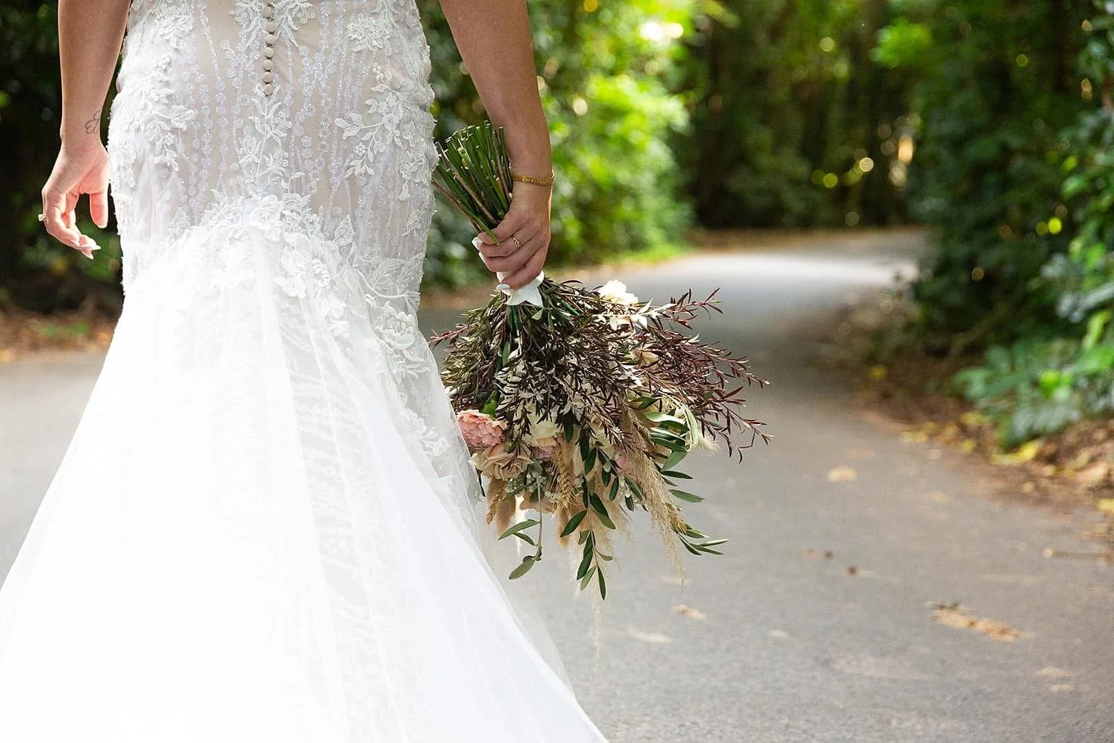 Close-up of a bride in a white wedding dress holding a bouquet of flowers on a pathway surrounded by green foliage.
